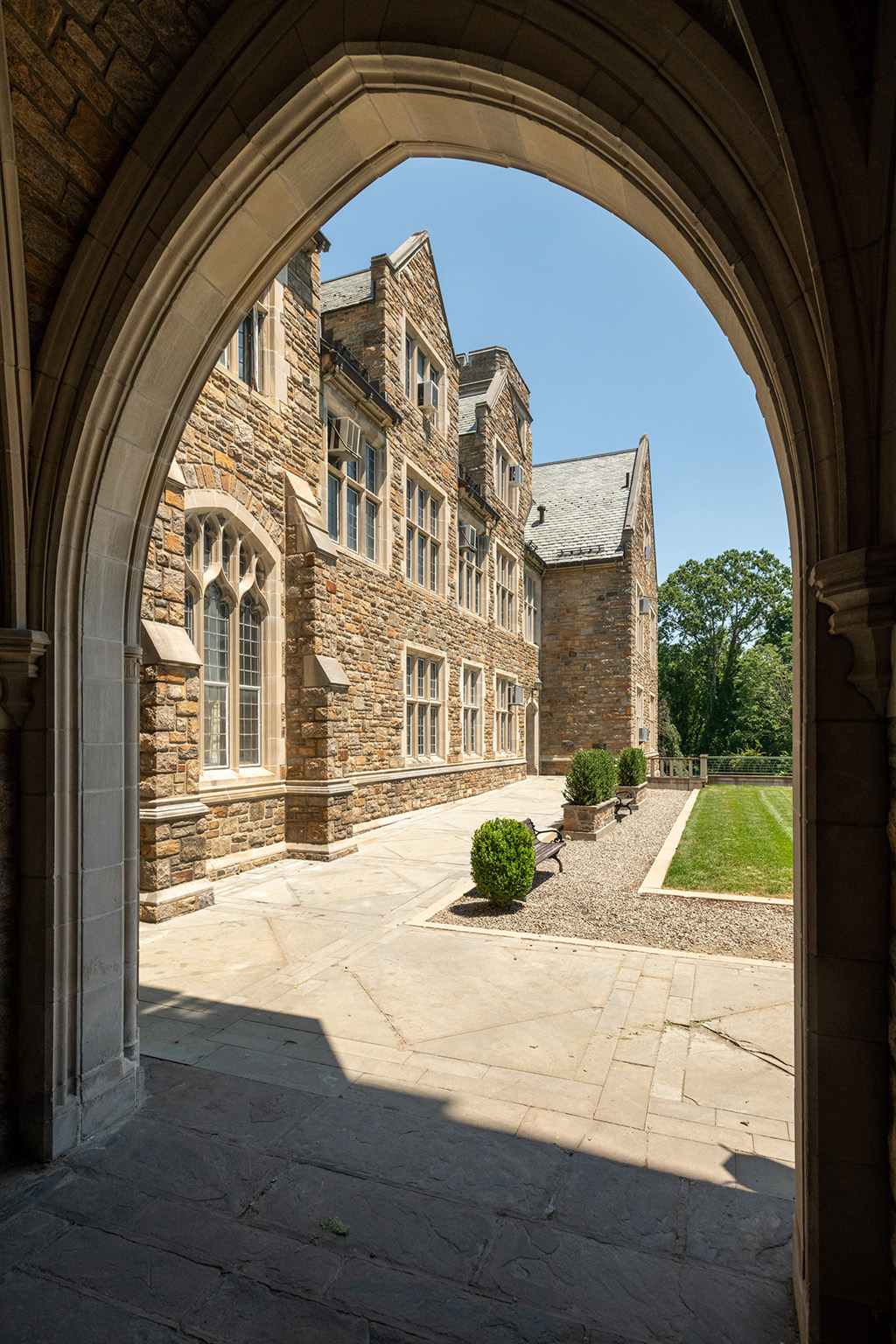 View through a stone archway framing a courtyard with neatly trimmed shrubs, benches, and the side of a large, multi-story stone building with tall, leaded-glass windows under a clear blue sky.