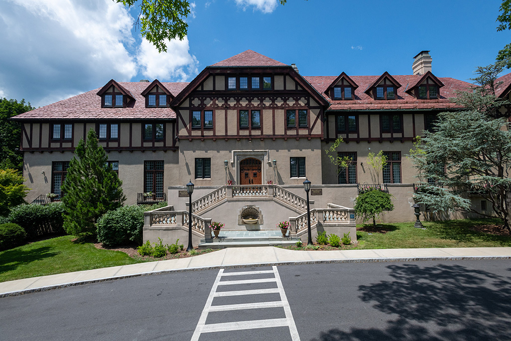 Front view of a large Tudor-style building with a red-tiled roof, decorative half-timbering, and a central stone staircase leading to a wooden double door entrance, flanked by landscaped greenery.
