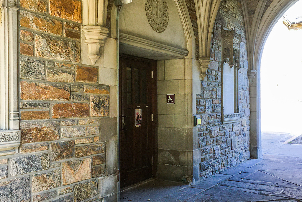 Stone archway entrance to a historic building, with a wooden door marked by an accessibility sign and surrounded by detailed stonework.
