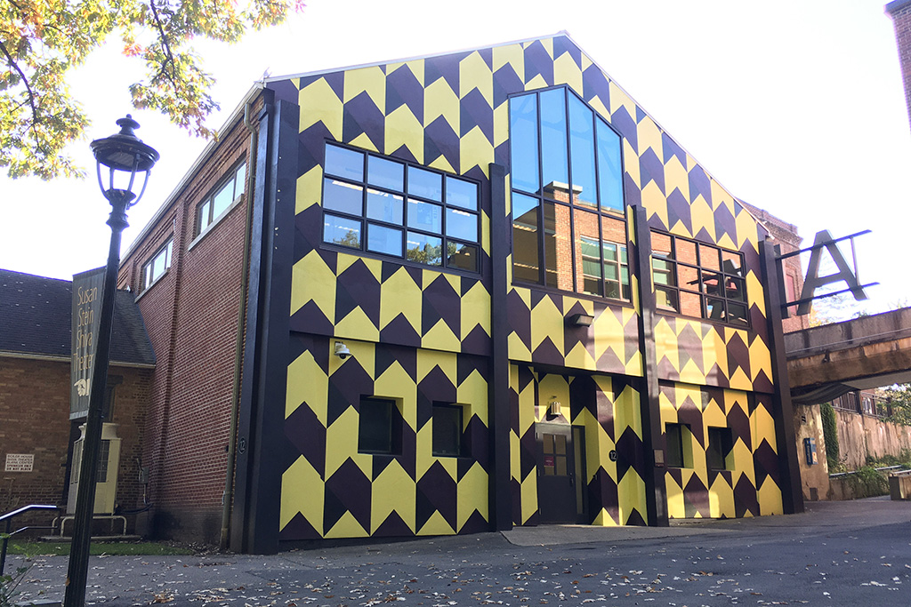 The full exterior of the building as viewed facing south from in front of the lower level. The face of the building is painted in a large-scale yellow and black offset chevron pattern.