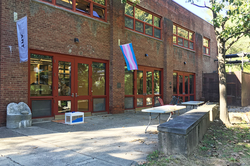 Exterior of a brick building with large red-framed windows and doors, displaying a white flag with the word 'ART' and a transgender pride flag. Several folding tables and benches are placed outside on a concrete patio, with a bicycle leaning against the wall and trees casting partial shade.