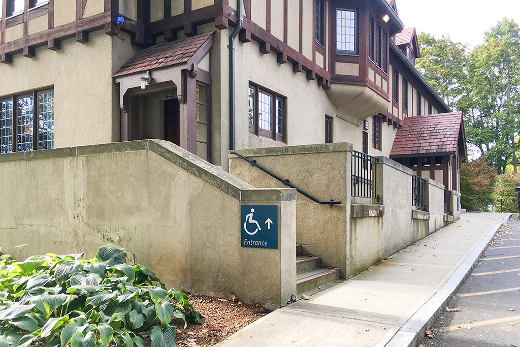 Accessible entrance ramp and stairs with handrails leading to a beige stucco and brown timber-framed building, with a sign indicating wheelchair access.