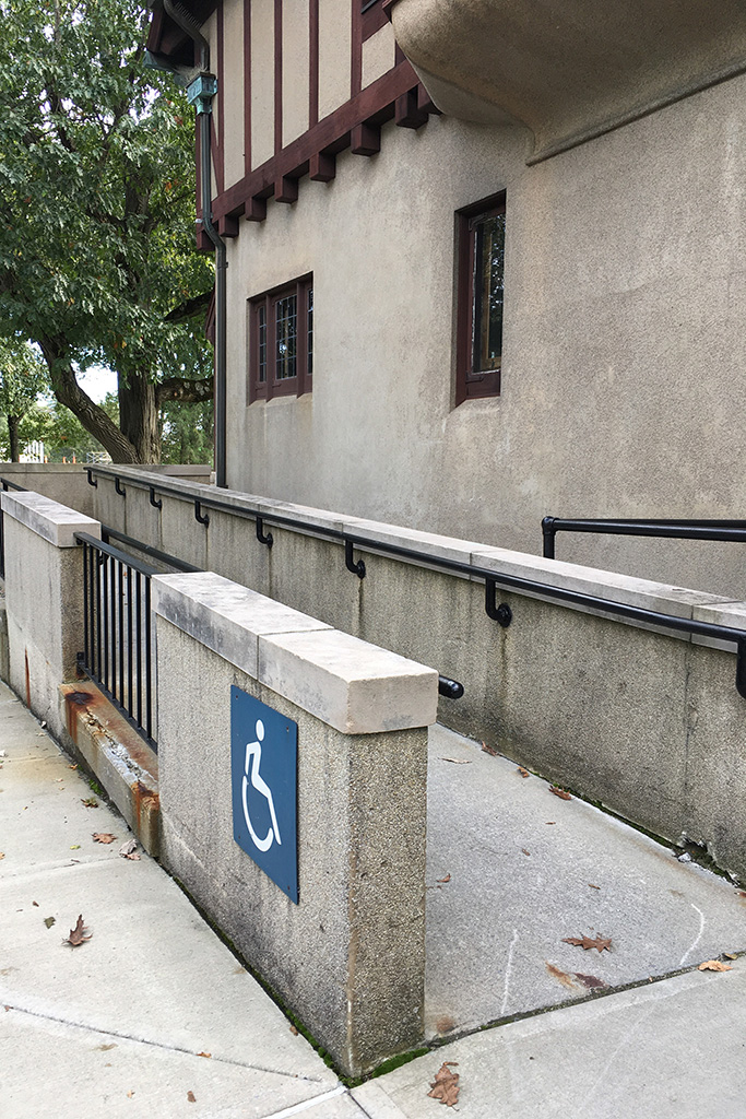 Concrete wheelchair-accessible ramp with black railings leading to a beige stucco building with red trim and small windows, shaded by a large tree.