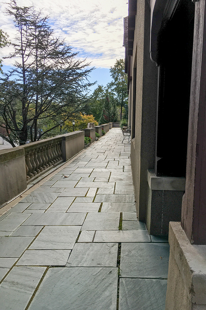 Narrow stone-tiled terrace alongside a building, bordered by a balustrade with views of trees under a partly cloudy sky.