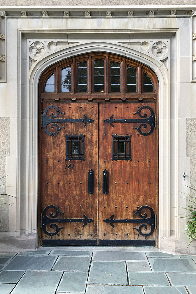 Ornate wooden double doors with decorative black wrought iron hinges and small square windows, set within a carved stone Gothic-style archway.