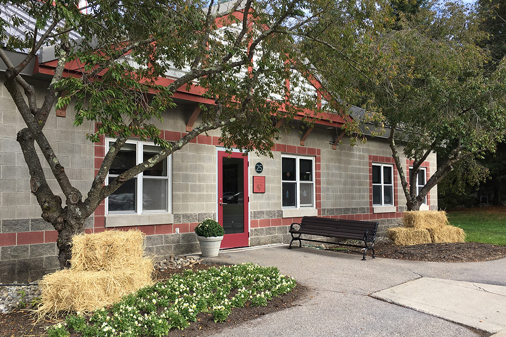 Exterior view of a single-story building with gray concrete block walls, red trim, and a red door, surrounded by trees and decorated with bales of hay and planters, with a bench placed near the entrance.