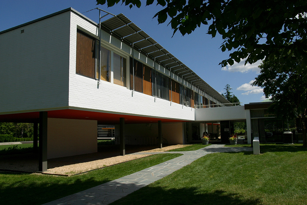 Modern two-story building with white brick exterior, large upper-level windows, and an overhanging design supported by columns, with a shaded walkway and landscaped lawn in the foreground under a sunny sky.