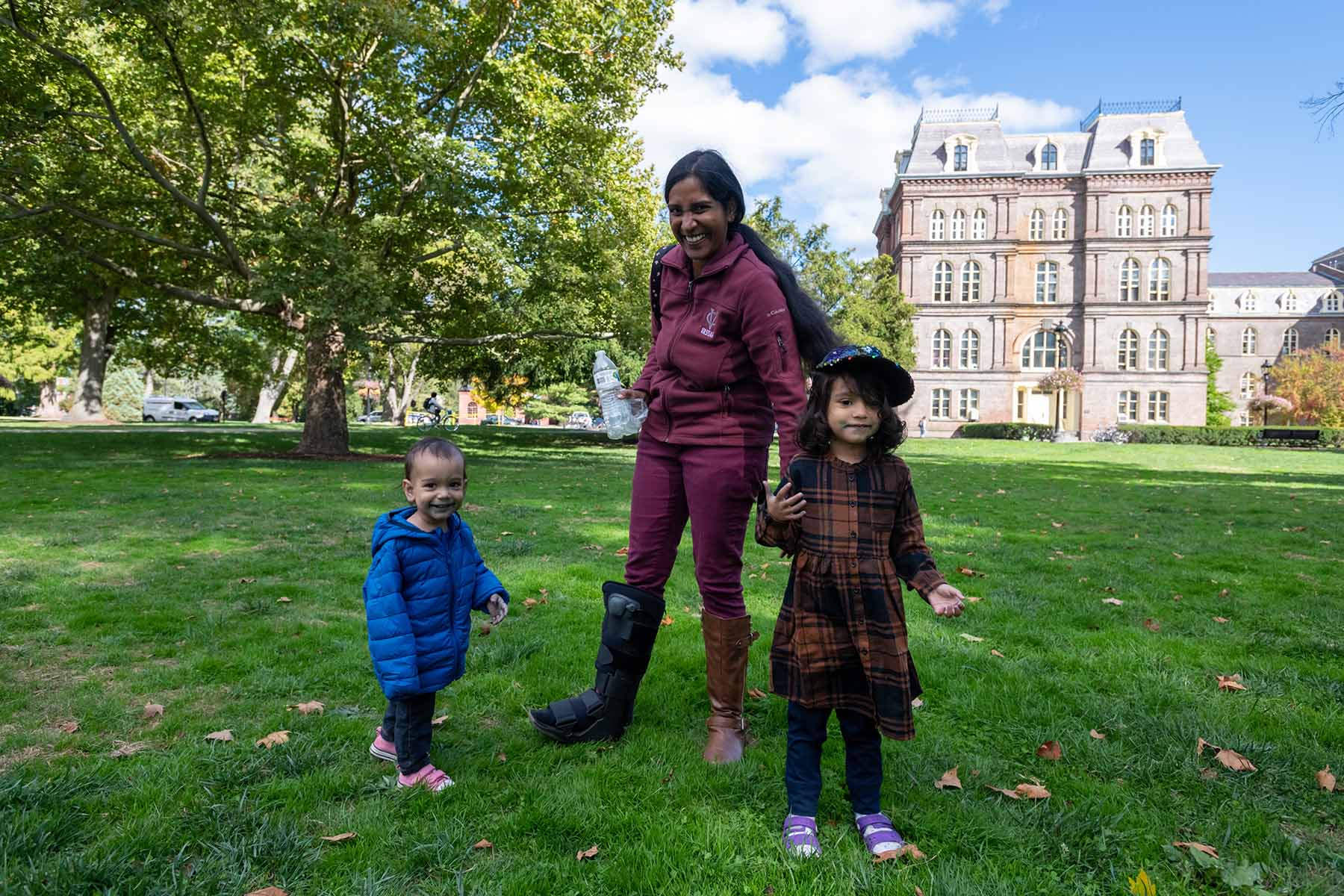 Woman with black cast on right foot/leg standing on lawn with 2 kids with Main Building in the background