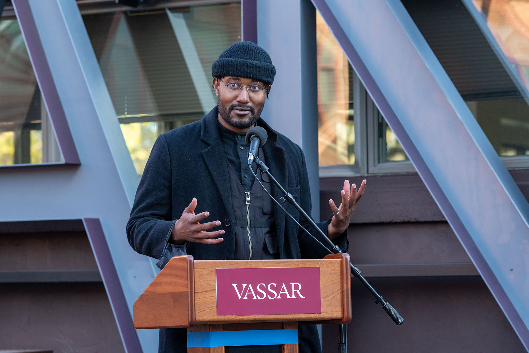 Man wearing a black shirt, black coat and black wool hat standing at a podium with "Vassar" on the front and microphone attached