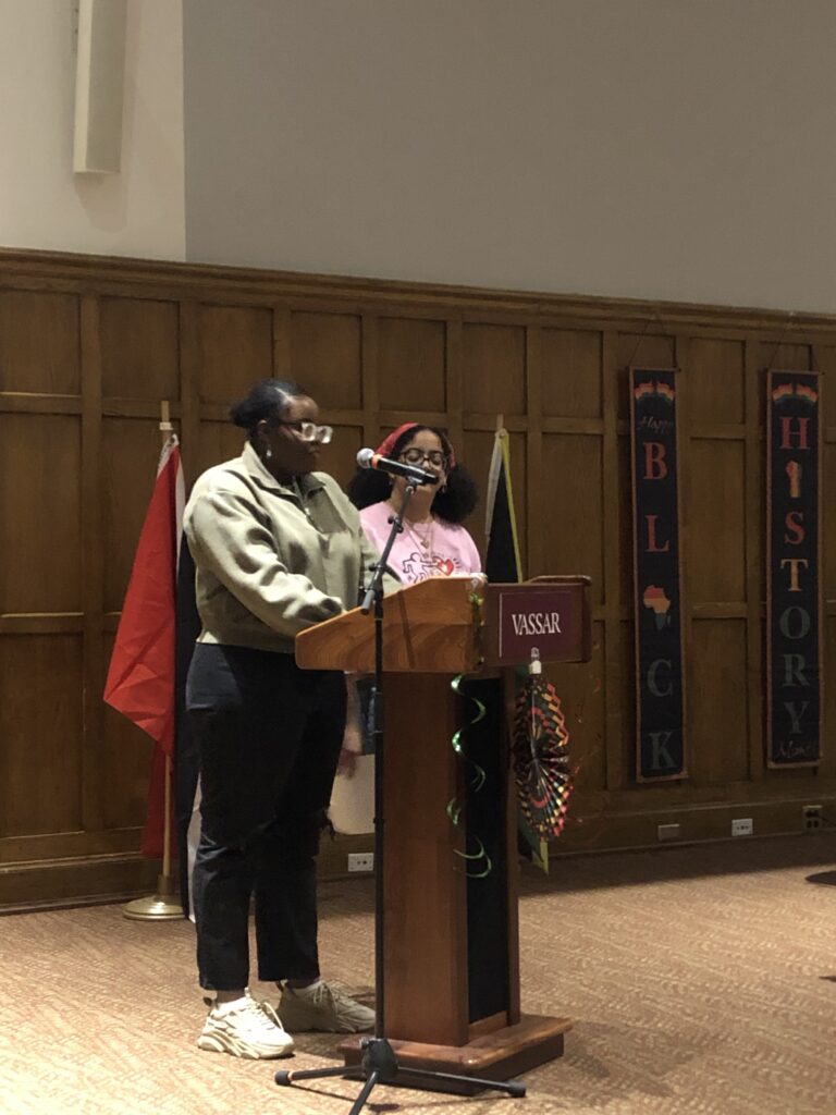 Students moderators Naomi Young and  Pierangelis Valerio standing at the podium speaking into the mic while addressing the audience. 