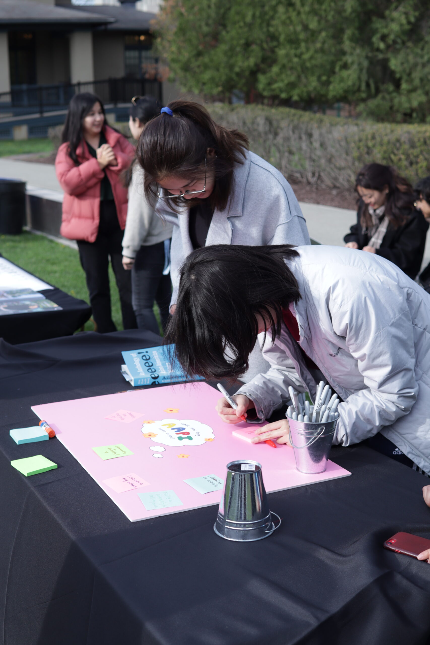 Two students with their heads down; one student is looking at the pink "What does being AAPI mean to you?" poster, while the other student is writing on a post it.  