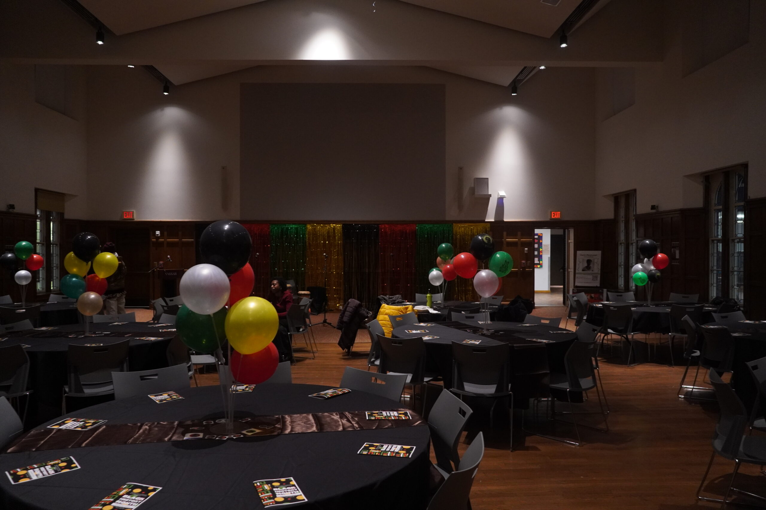 Round tables with black table cloths and brown runners. Different colors of green, white, orange, red, yellow and black balloons photographed as table centerpieces.