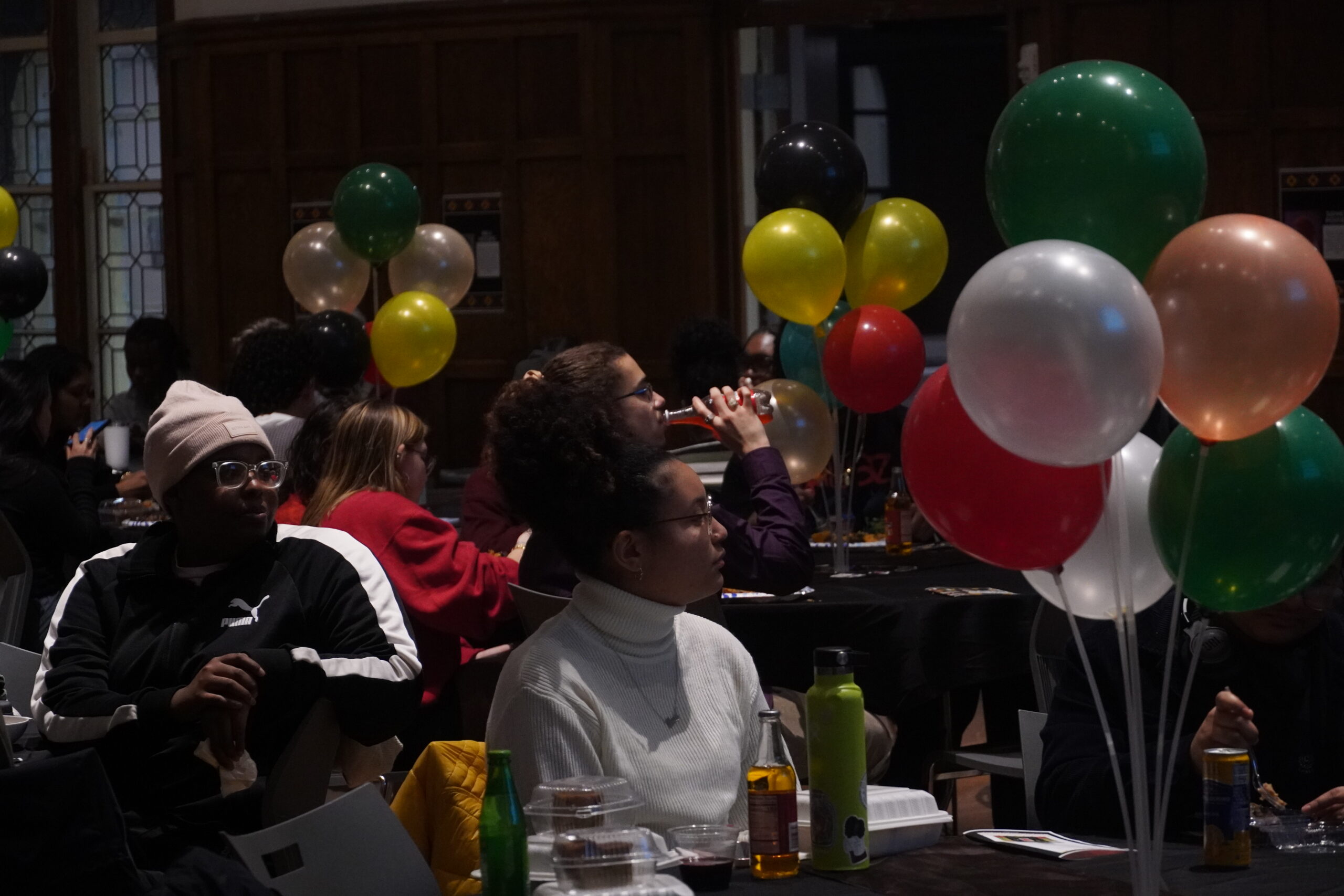 Students seated at tables watching performances take place at the kickoff. Different colors of green, white, orange, red, yellow and black balloons photographed as table centerpieces.