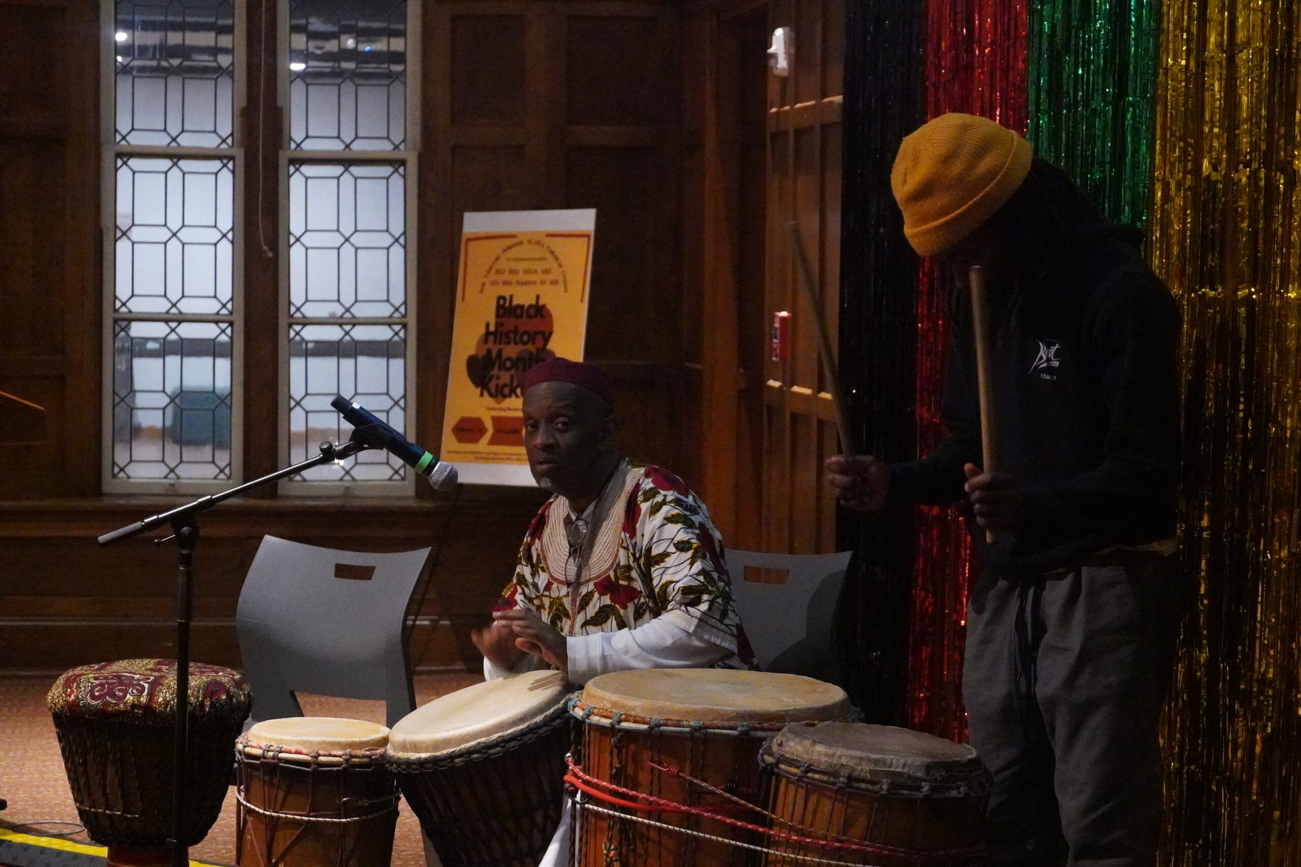 Baba Kazi Oliver seated with drums three drums in front of him and Nick Jackson standing to his left side playing the two drums in front of him.