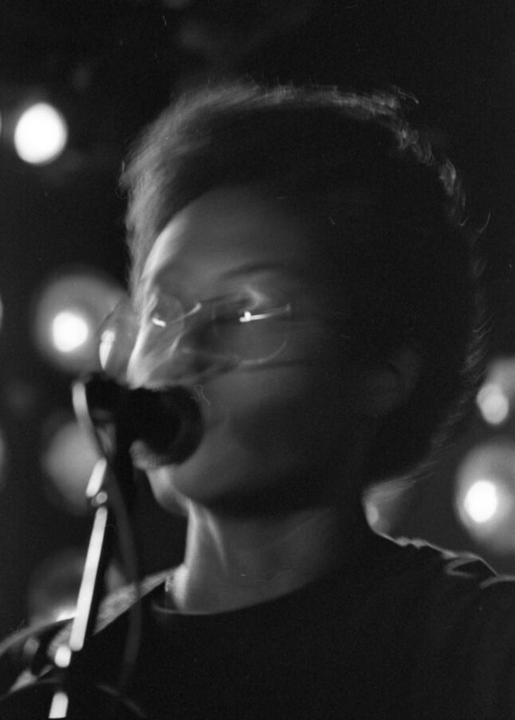 Black and white photo of artist tofusmell, or Rae Chen, at a concert singing in to the microphone. 