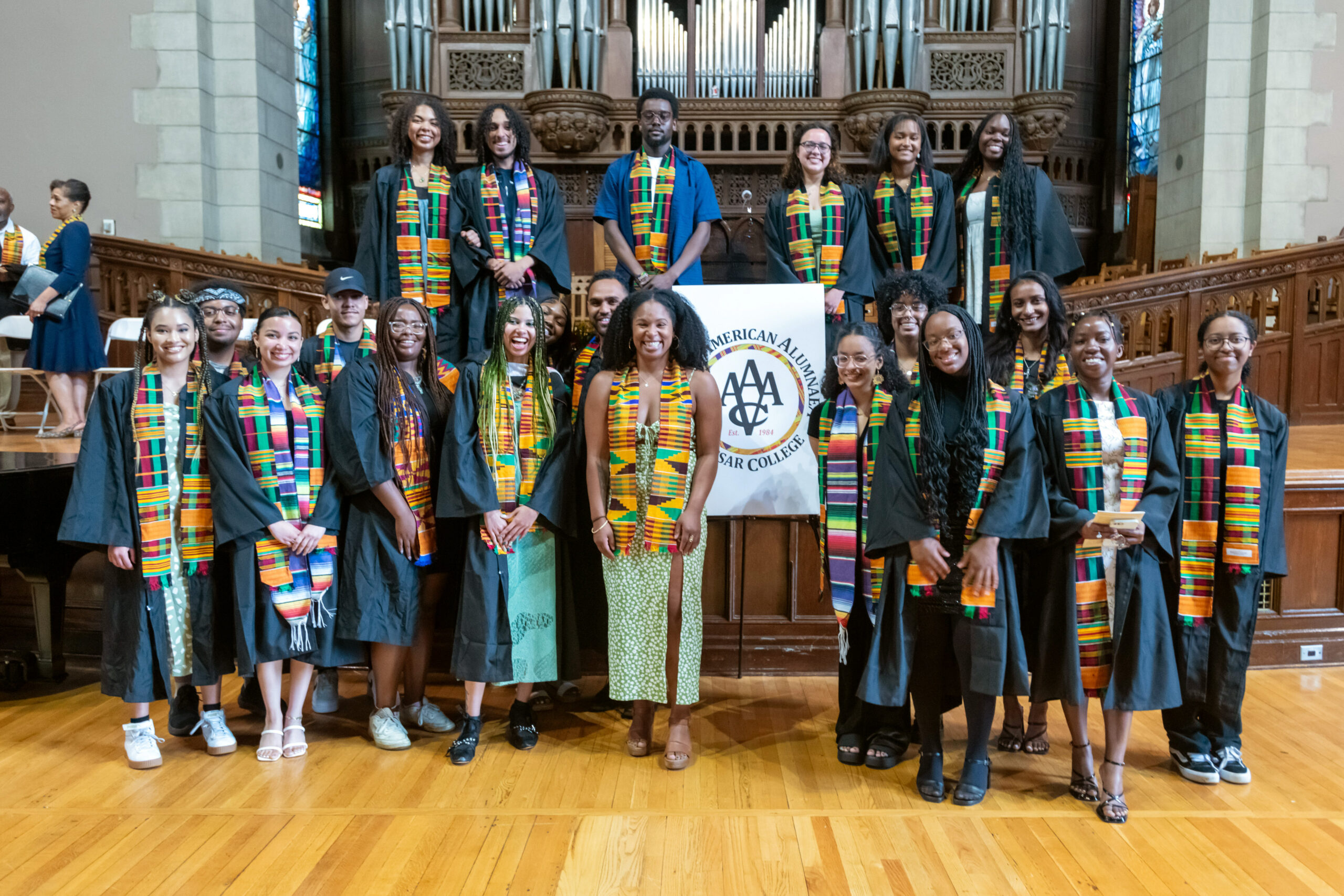 Twenty-One students standing at the Center of the Chapel (six students are standing on the chapel stage while fifteen are standing below the stage). The students are standing in front of the "African American Alumnae/i of Vassar College (AAAVC) logo displayed on an easel located in the center of the group photo.  