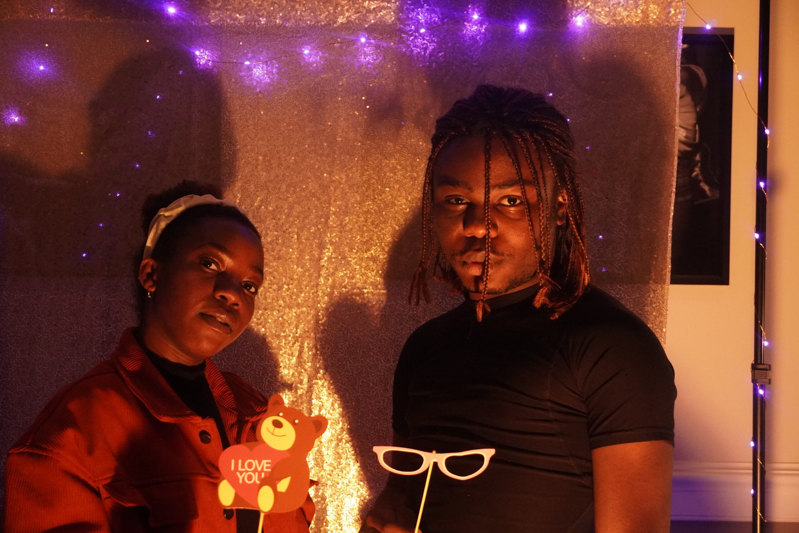 Two students, Bre and Favour standing inf front of the golden sequenced backdrop with blue lights across the top, as they pose for the camera with valentine's themed props.