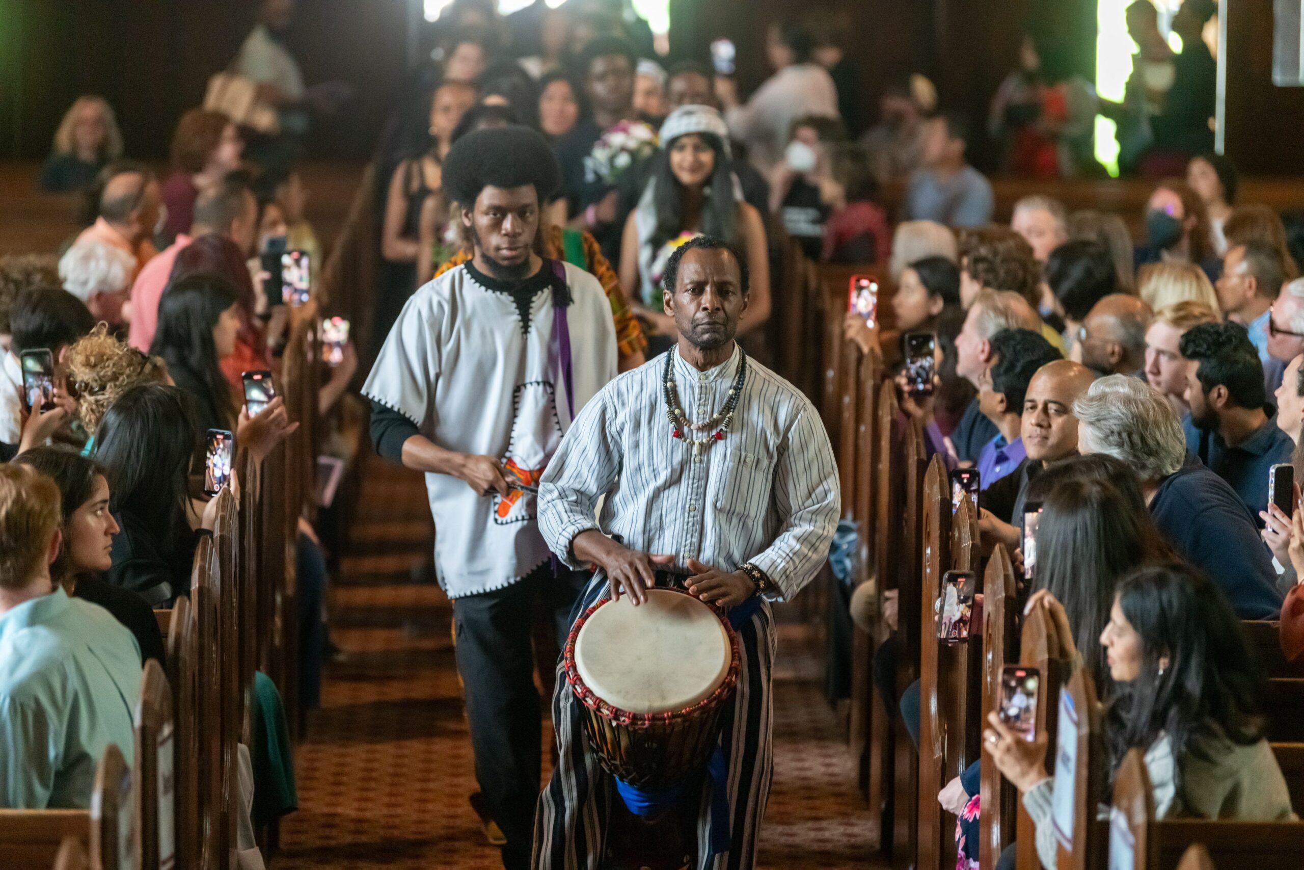 Karimu Drummers leading the procession. Two of the drummers are walking down the Chapel aisle playing the drums with African Violets walking behind them as attendees are seated on both sides of the chapel.