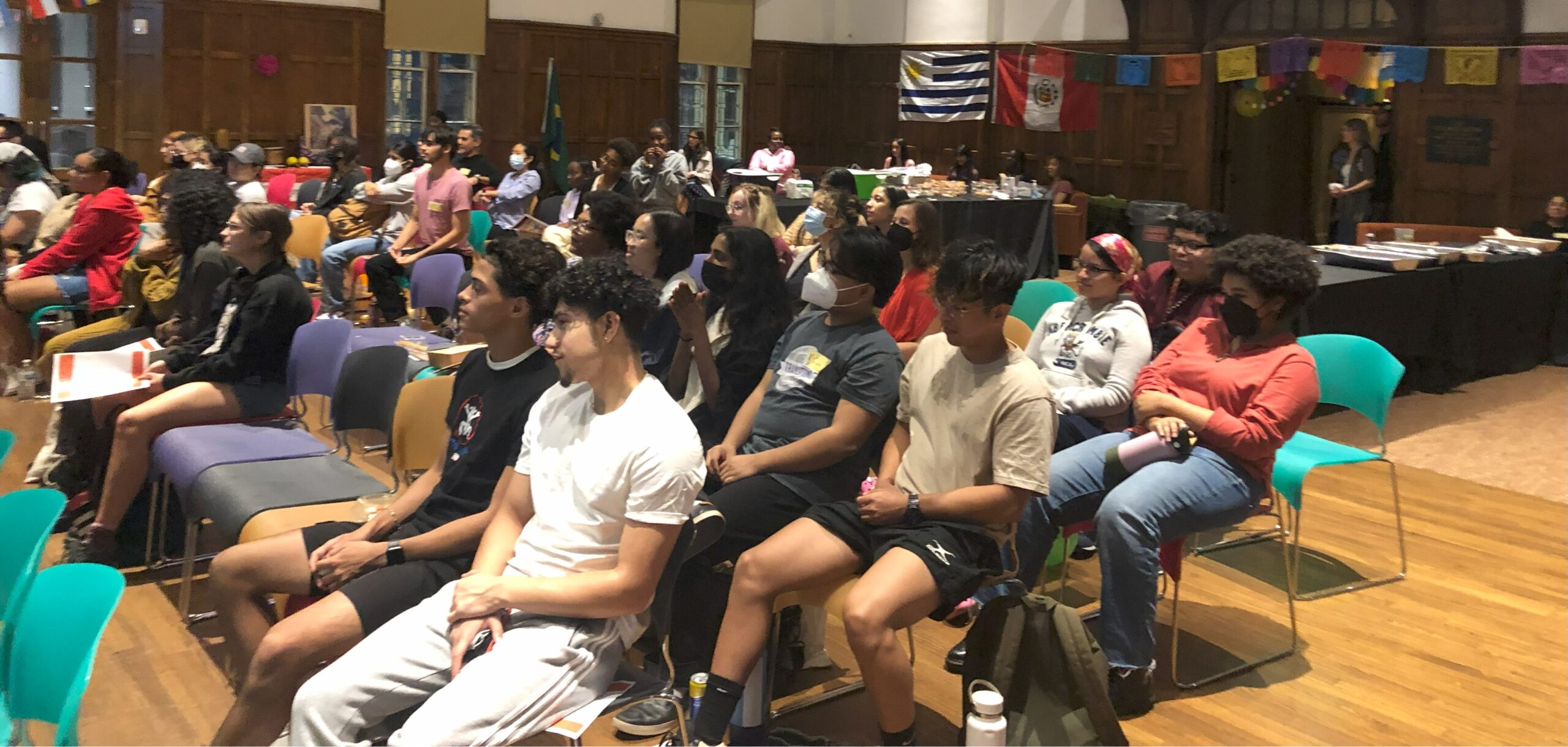 Large group of students sitting next to each other in multicolored chairs, some with masks, some unmasked as they watch the program.