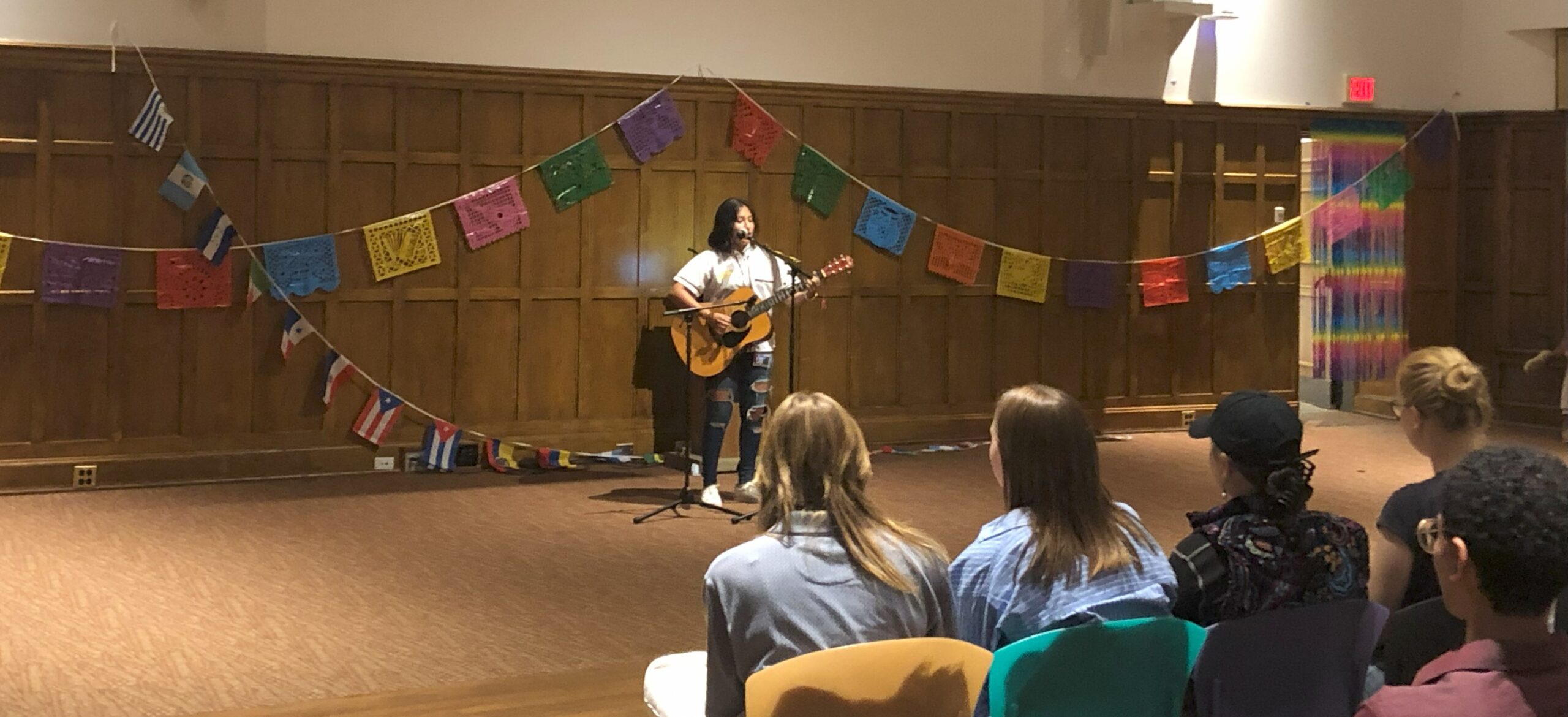 Darianna standing at the center of the stage, behind two mic stands with one mic positioned at her wooden guitar and the other mic positioned near her face as she sings and plays an acoustic song.
