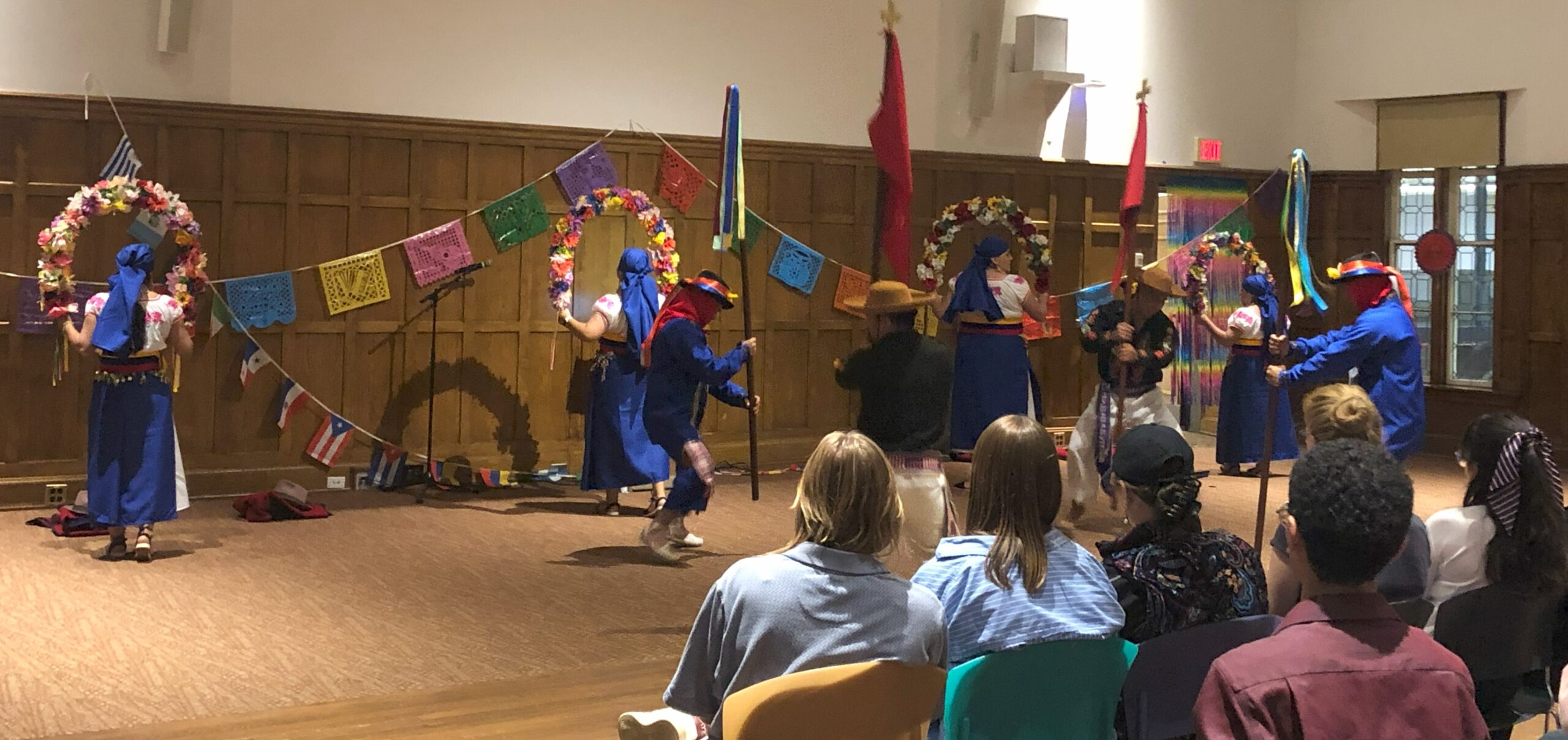 Dance group with eight members pictured standing on stage. The women have their backs towards the audience holding large flower wreaths in their hands while wearing blue scarfs, white with pink flower print shirts and blue long skirts. Two men are holding poles with yellow/blue decor, blue shirts and black hats with red and yellow decor. Two men are holding poles with a red flag and gold cross at the top while wearing black shirts, white pants and tan hats. 