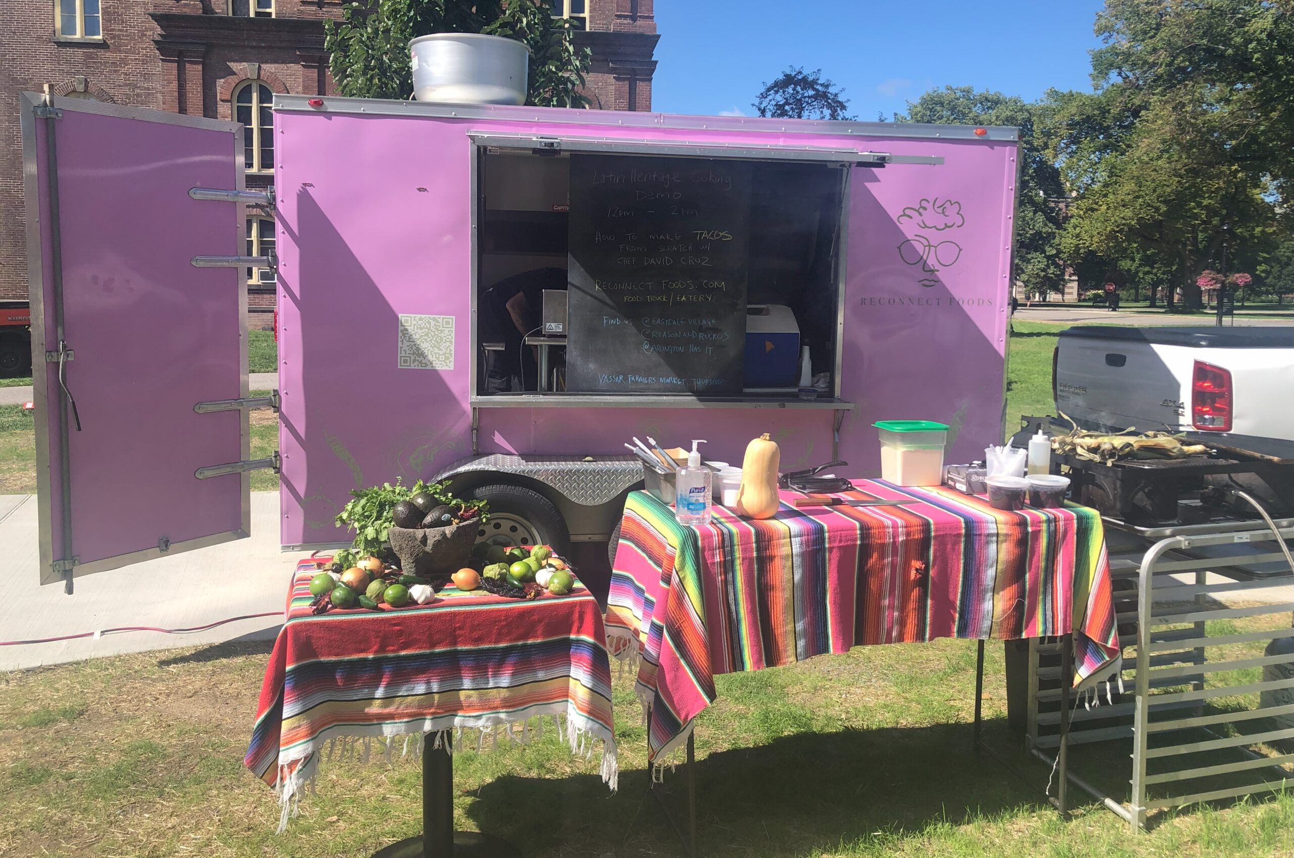 Pink  food truck with it's back doors open, window open with a large chalk board that's titled. "Latin Heritage Cooking Demo" along with menu, social media info and two tables with Latin patterned table cloths and fruits, vegetables, cooking tools. 