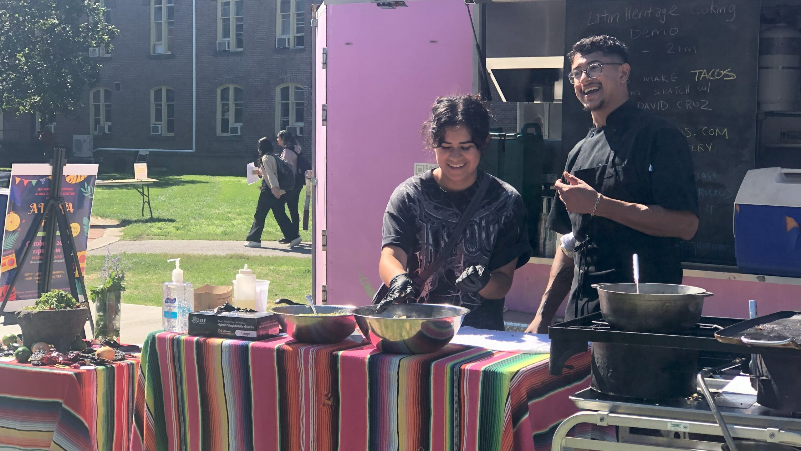 Student standing next to Chef Cruz as she grabs the large metal bowl Chef smiles holding his left arm up and looks into the crowd.