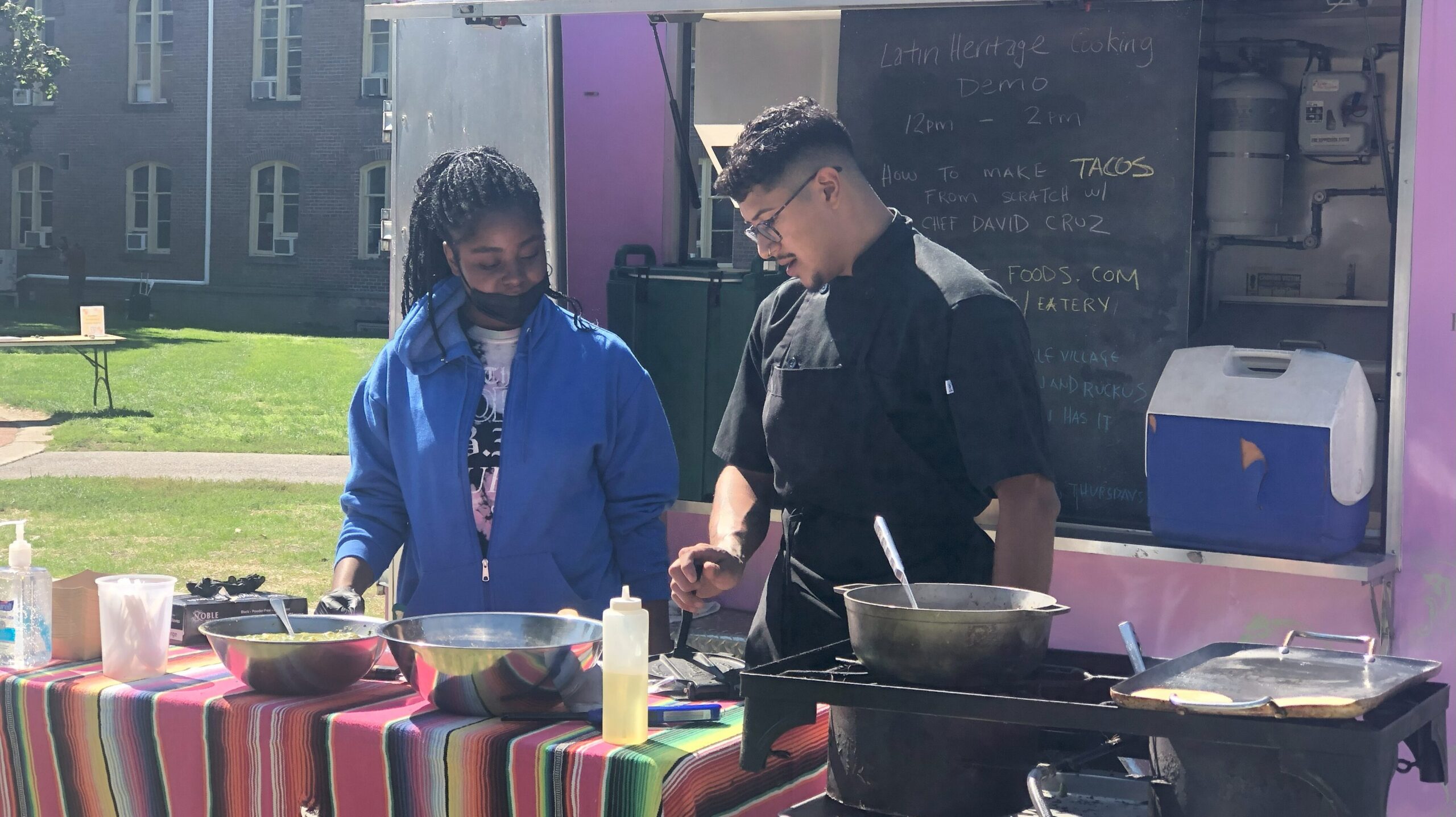 Student and Chef Cruz standing next to each other behind the table. The student is looks down at Chef's hand motions while he looks down and explains the next step.