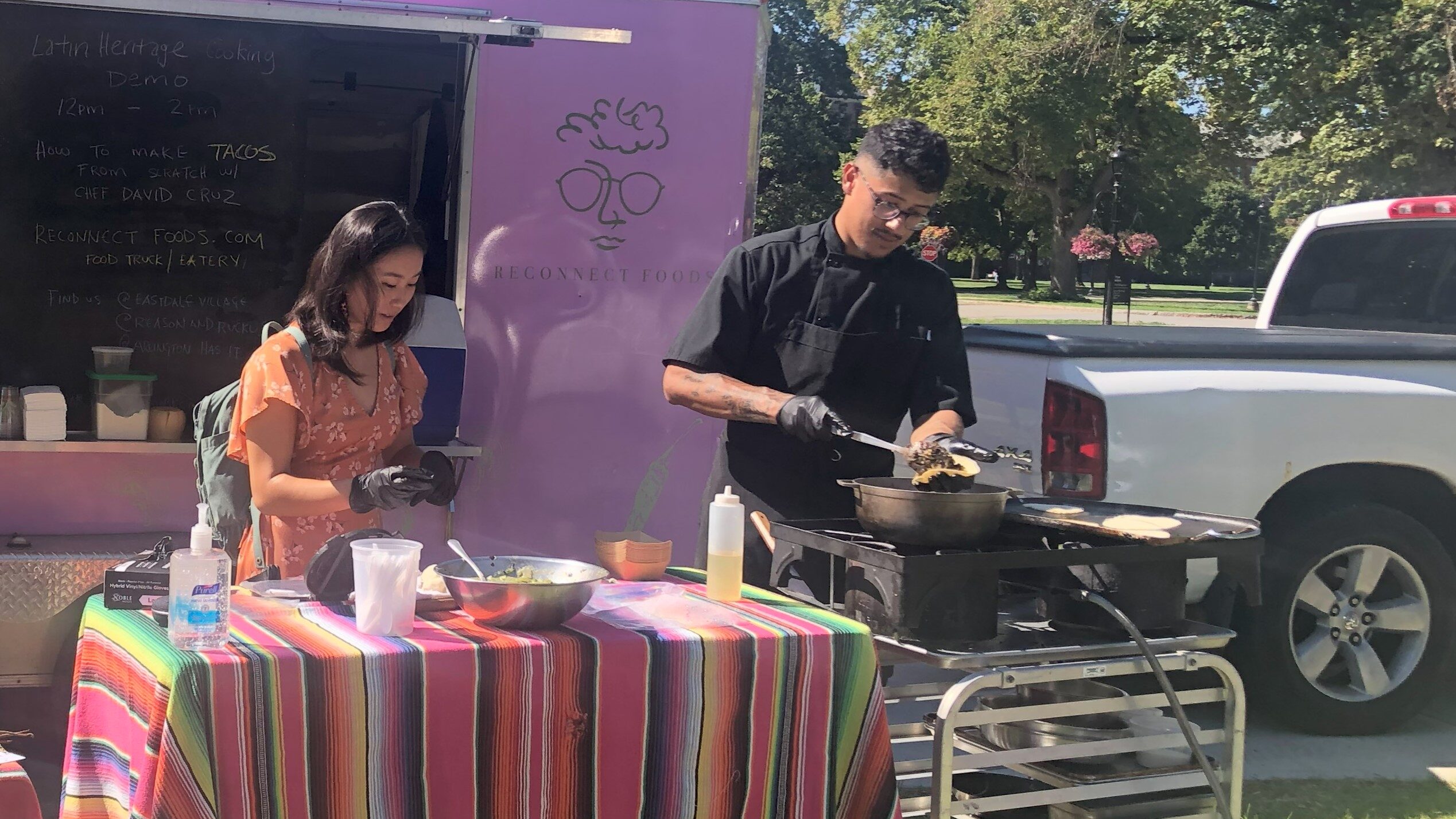 Student wearing gloves standing in front of the table with Chef Cruz standing next to her cooking over an electric pan.