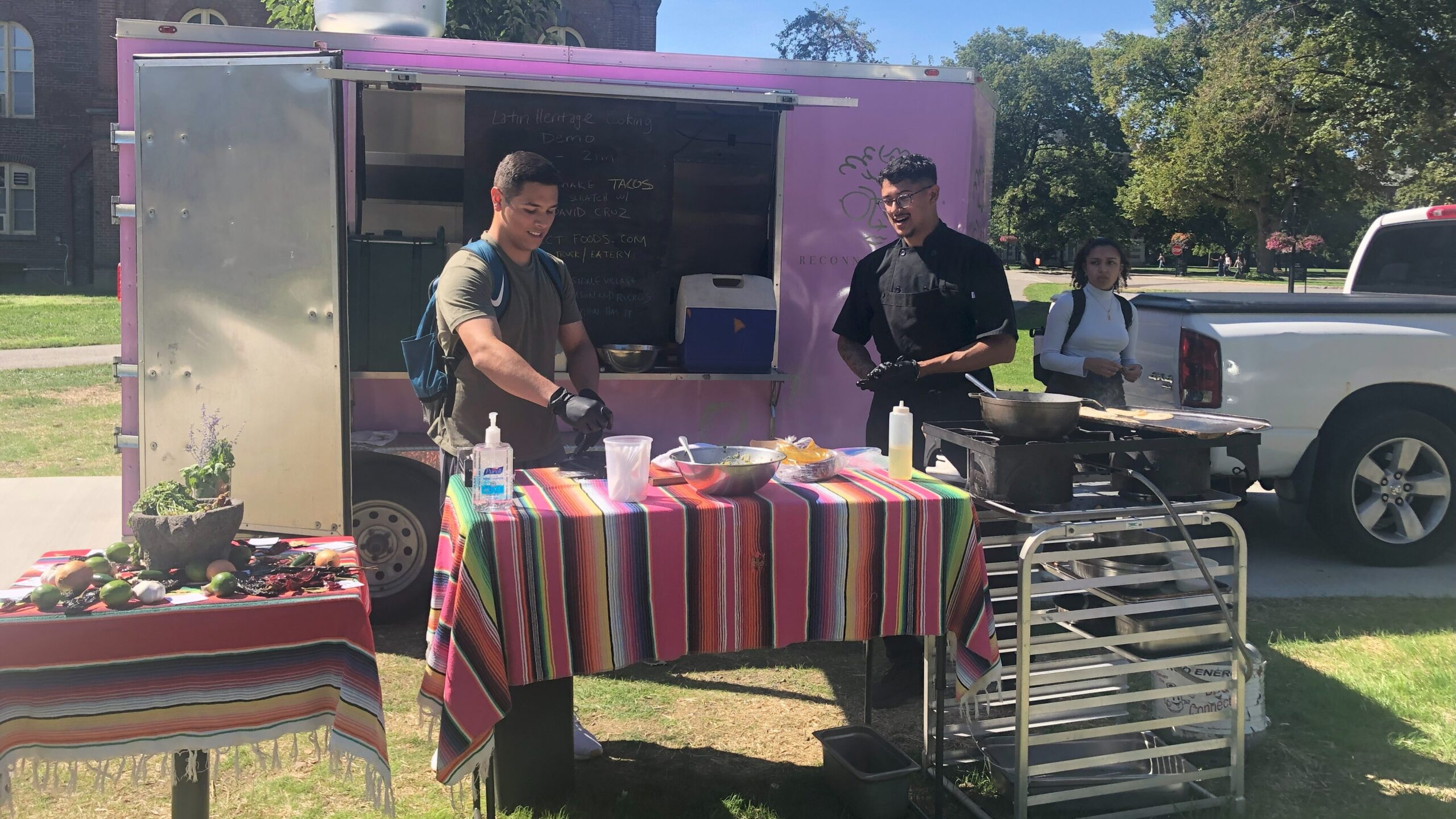 Student standing near Chef Cruz as the student holds an ingredient over the table and Chef looks on talking him through the next steps.