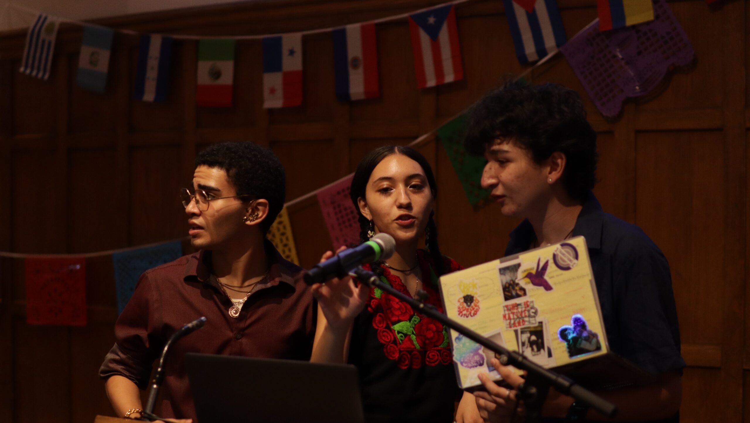 David, Sofia and Óscar standing side by side behind the podium, Sofia is touching the mic and speaking to Óscar while Óscar is looking at the mic and holding his laptop. David is looking off to his left.