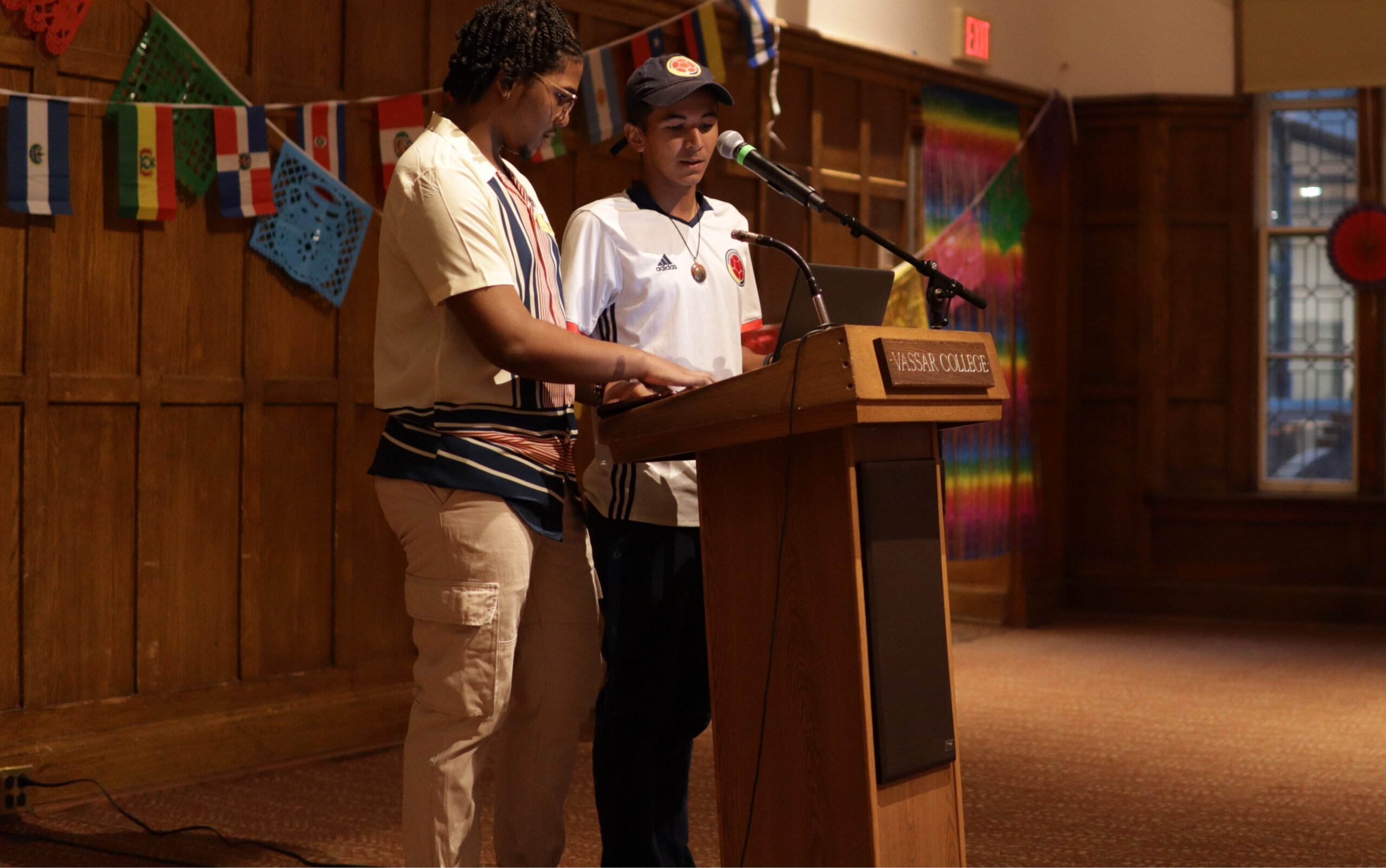 Carl and Mateo standing at the podium looking down with the mic in the center of them as various Latin country flags are displayed behind them.