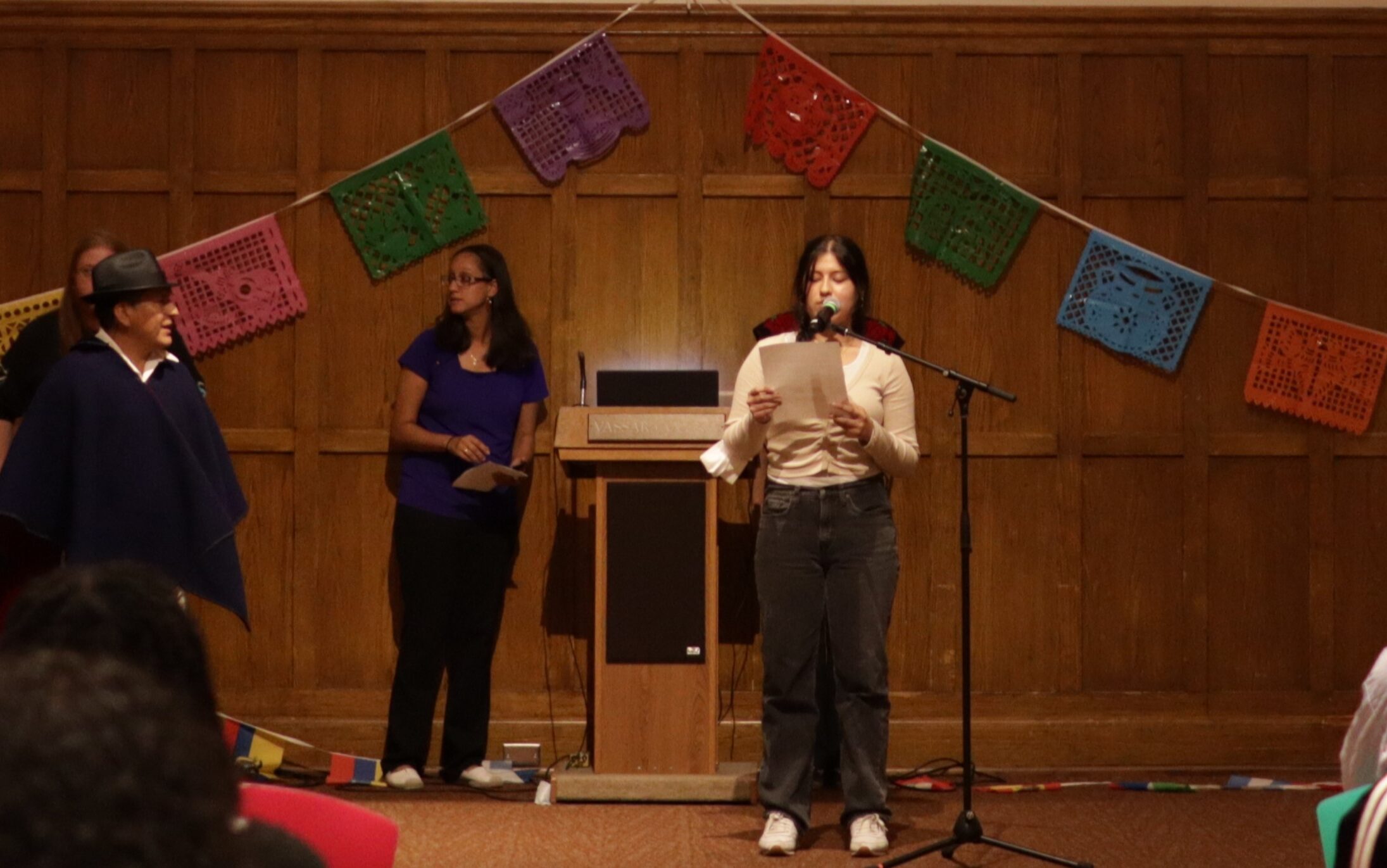 Sharon Llanes standing next to the mic stand reading from a paper that she is holding in front of her as she speaks into the mic.