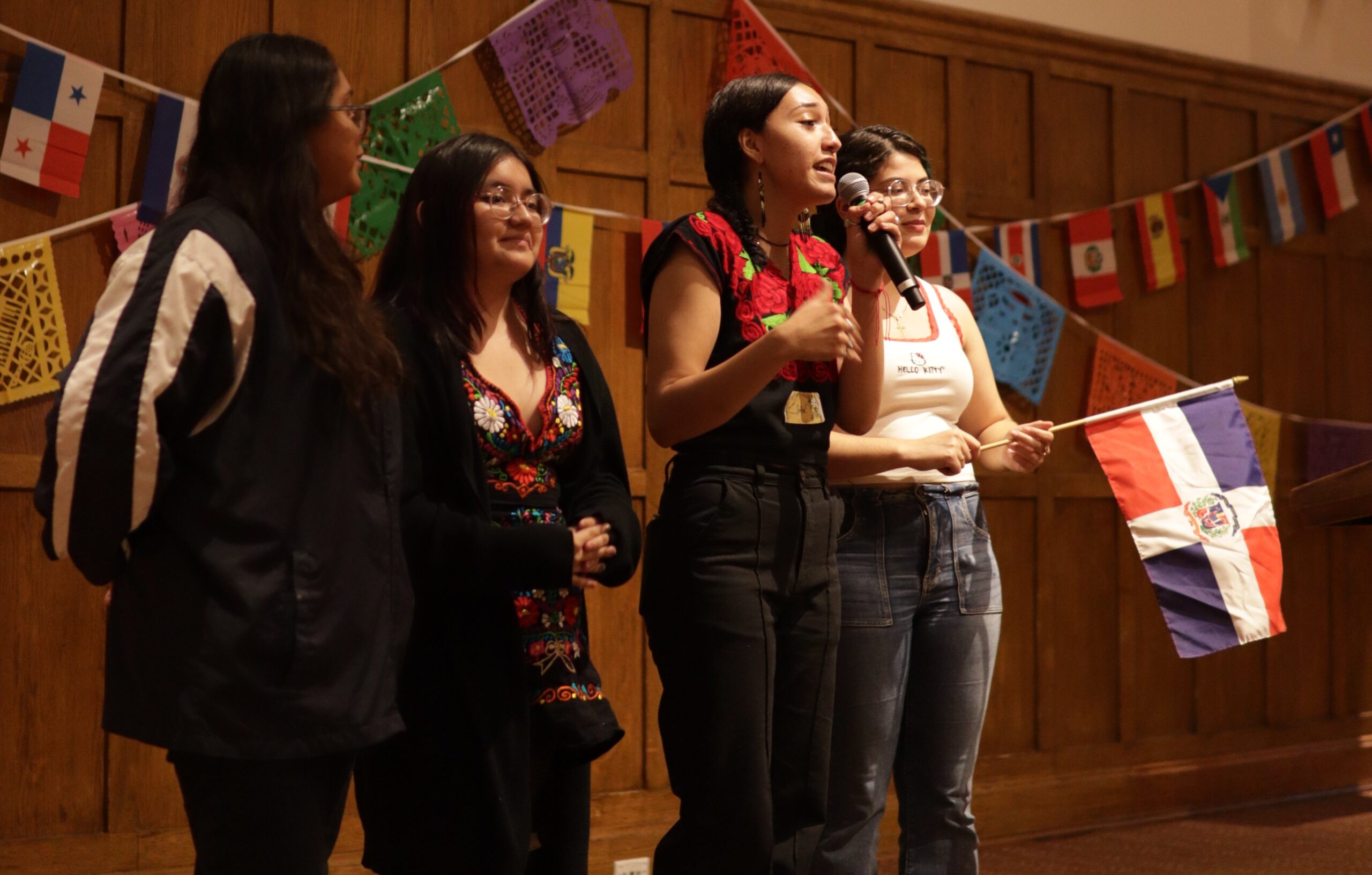 Four students standing side by side at the center of the stage. One student is speaking into the mic she is holding with her left hand.