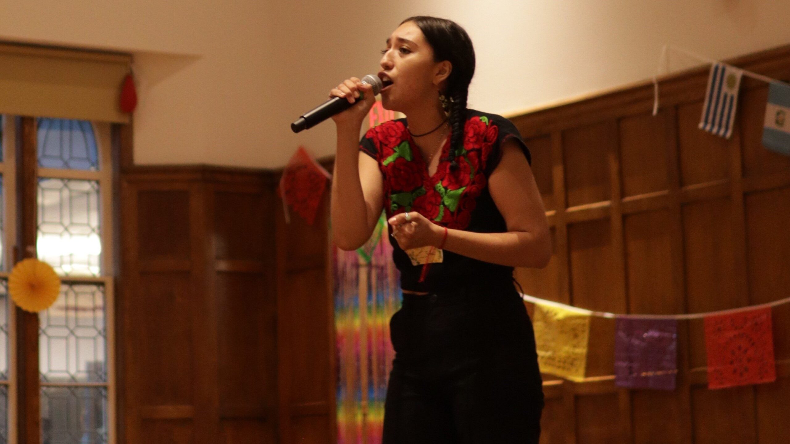 Student standing in the front of the Villard singing into a wireless mic that's held in her right hand, wearing a red and green floral design over her black shirt and wearing black pants.
