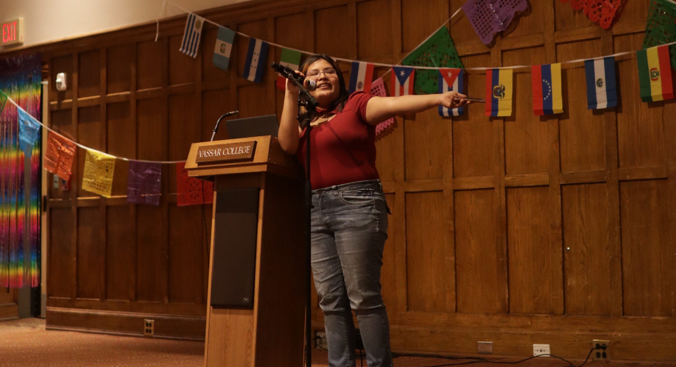 Melisa Torres standing at the center of the stage next to the podium with her right hand on the mic attached to the mic stand and her left arm extended out as she looks to her left.