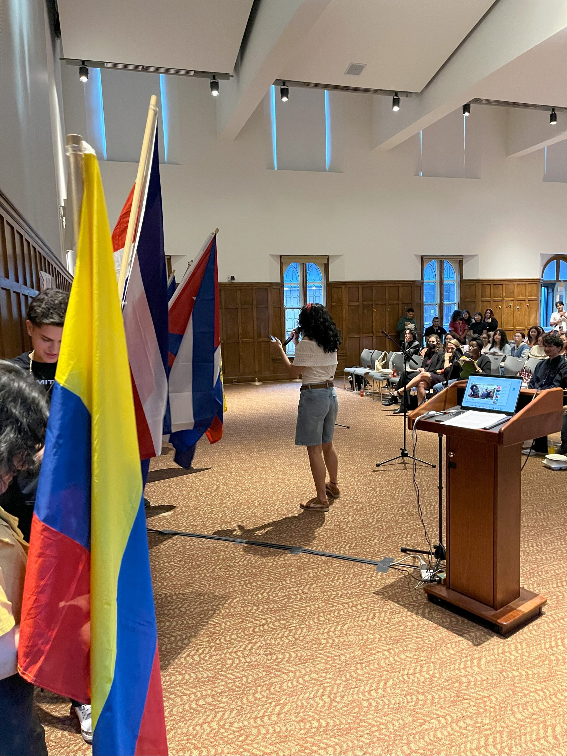 Sofia leading the "Que Vive" Ceremony as she stands in front holding a mic reading off a paper in her other hand while the flag holders are standing to the right lined up against the Villard wall.