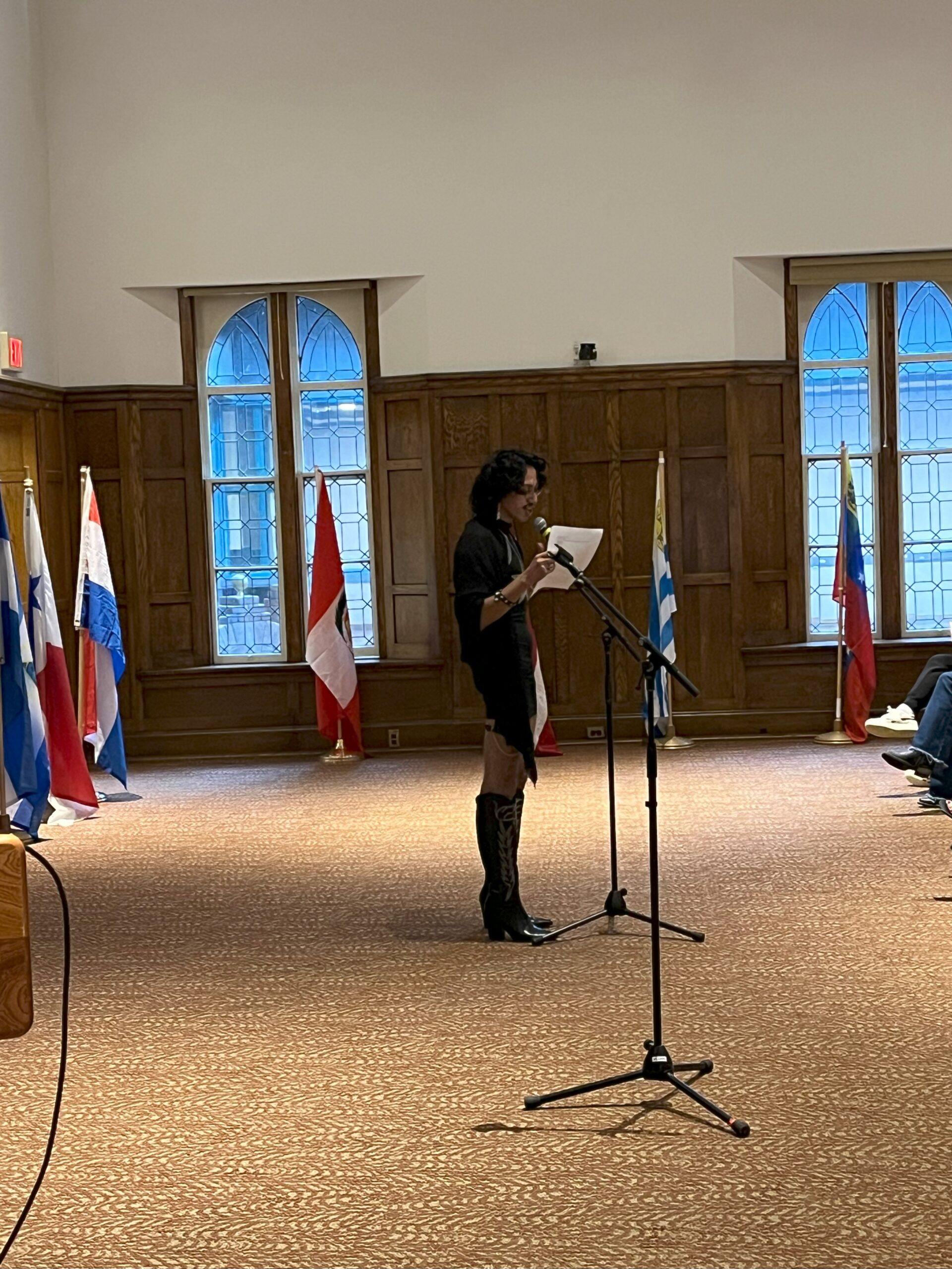 Alma standing at the front of the stage with a mic as they recite a poem they are reading off a paper. There are Latin American flags around the room in the back of Alma.