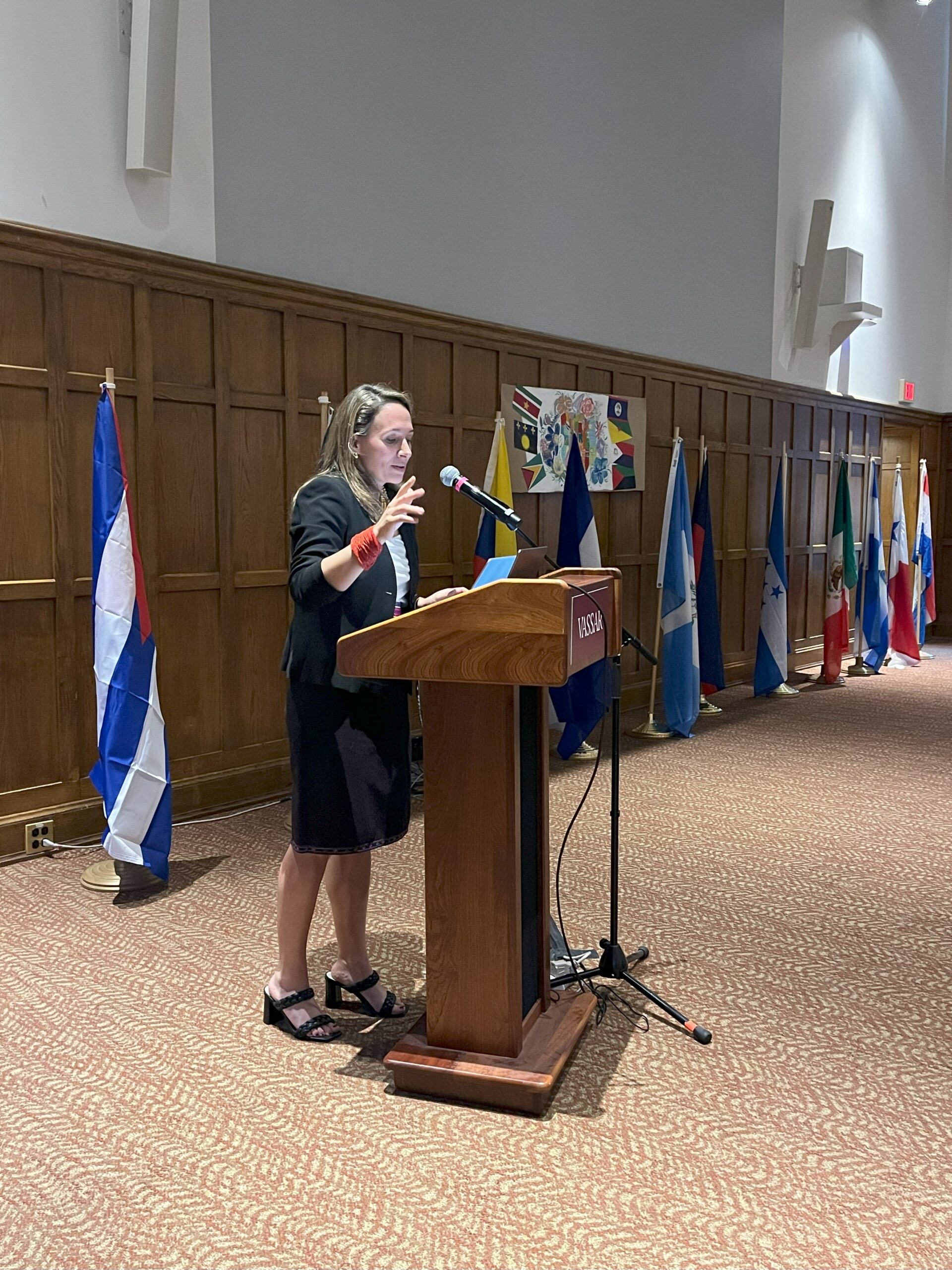 Natalia standing at the podium speaking into the mic as her right hand is lifted up. There are Latin American flags displayed in behind Natalia.