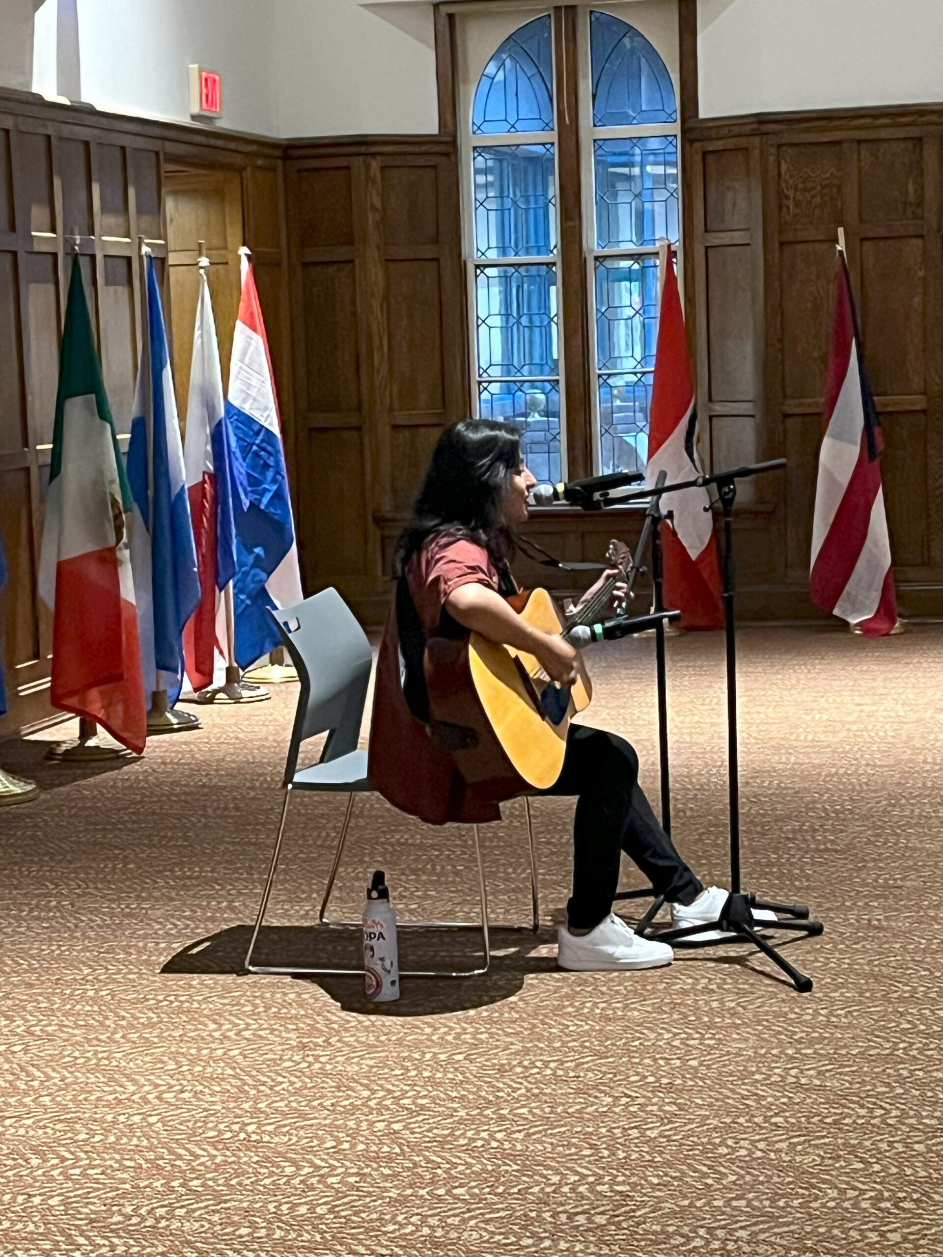 Darianna seated at the center of the stage with two mics in front of her. One mic she is singing into while the other mic is directed at her acoustic guitar.