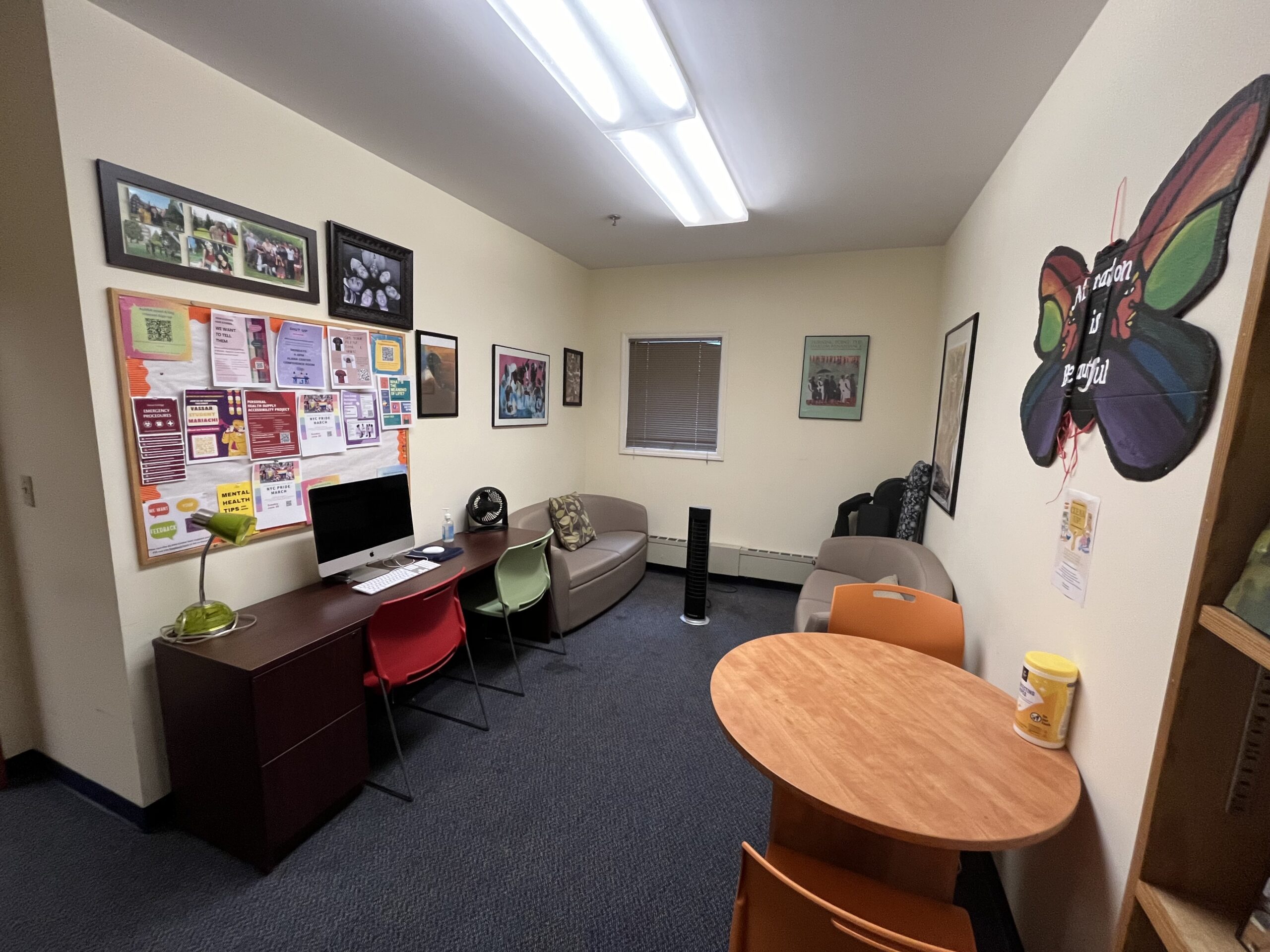 Round wooden table with two orange chairs on each side of the table, expresso colored desk with a silver mac desktop, green desk lamp, two chairs (red and green), two tan/green colored sofas on each side of the room with a standing fan in the center.
