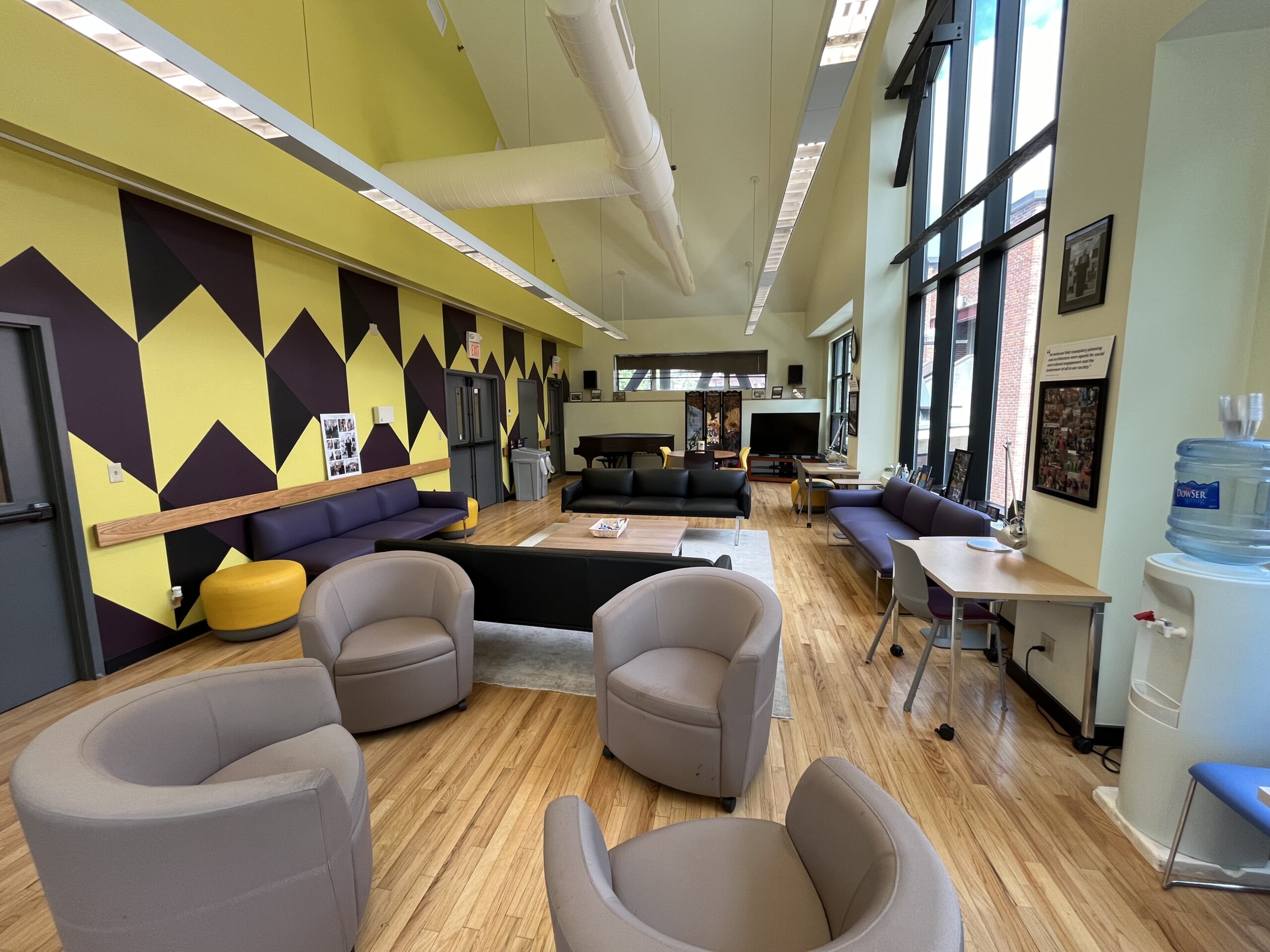 Community Room with four biege u-shaped swivel chairs, two black leather sofas, two purple leather sofas, two small desks with silver metal lamps, a wooden square center table and round table, piano and purple/yellow accent wall.