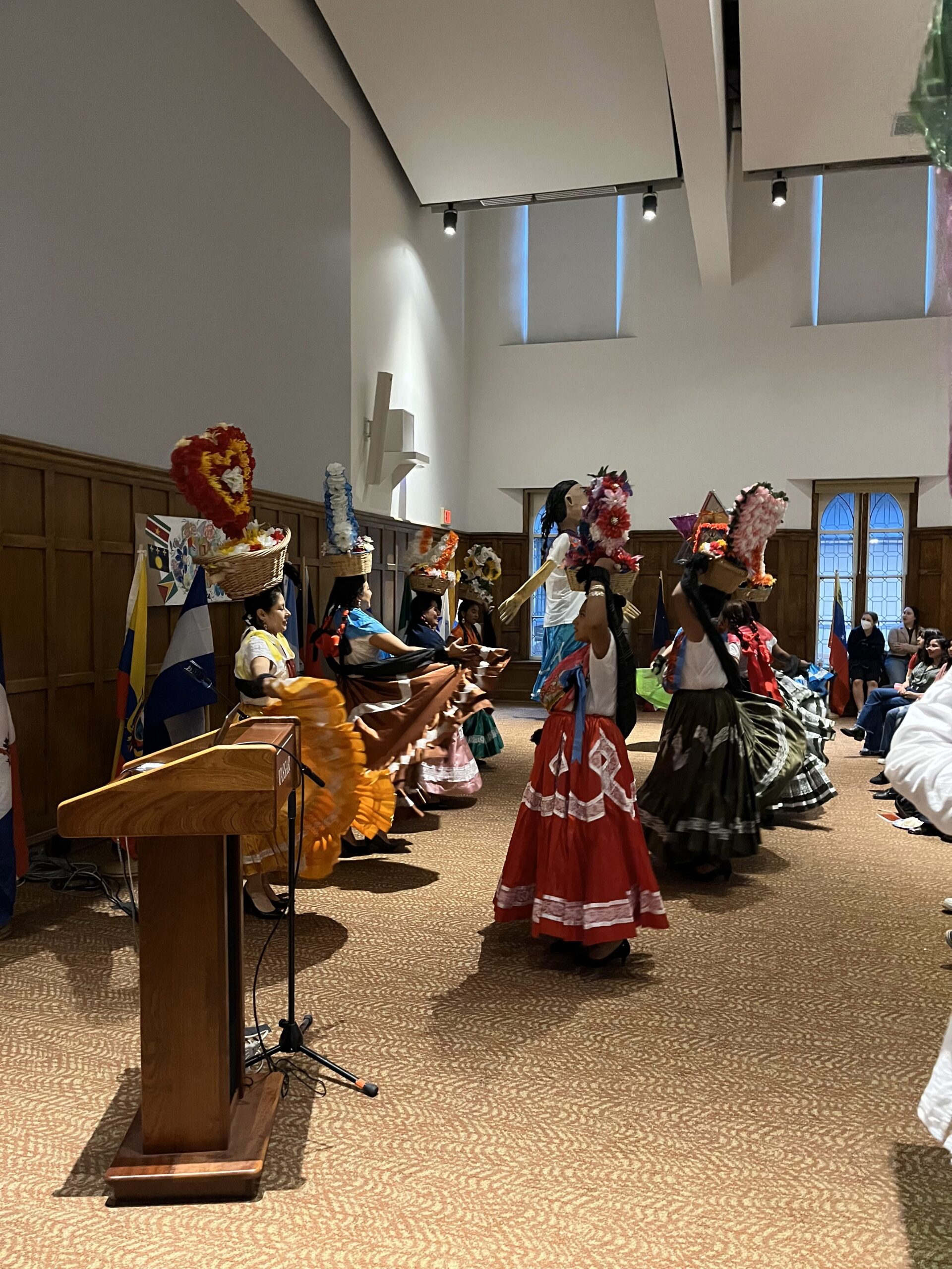 Women lined up side by side, facing one another while holding wooden baskets containing large floral arrangements on the tops of their heads as they perform a dance.