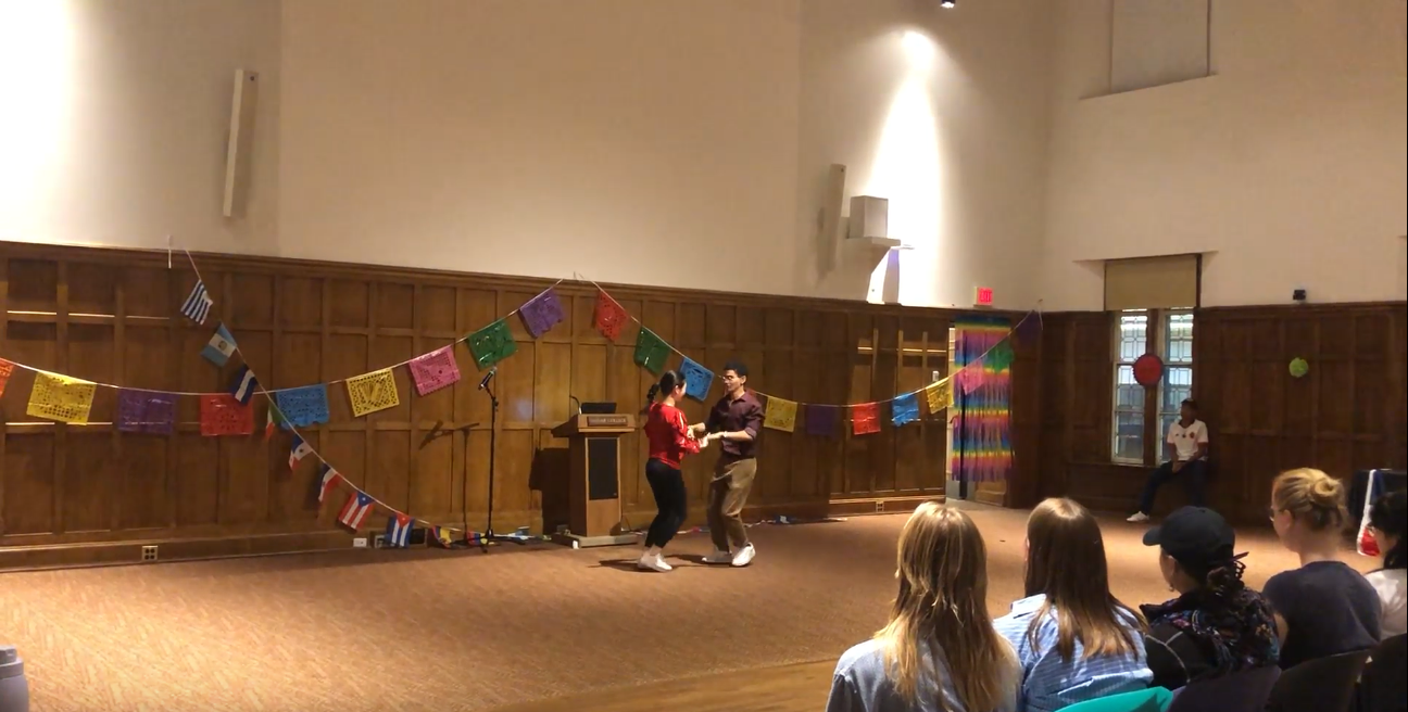 Students holding both hands as they perform a salsa dance. The student on the right is wearing a red shirt with black pants and the student on the right is wearing a dark down shirt with brown pants.