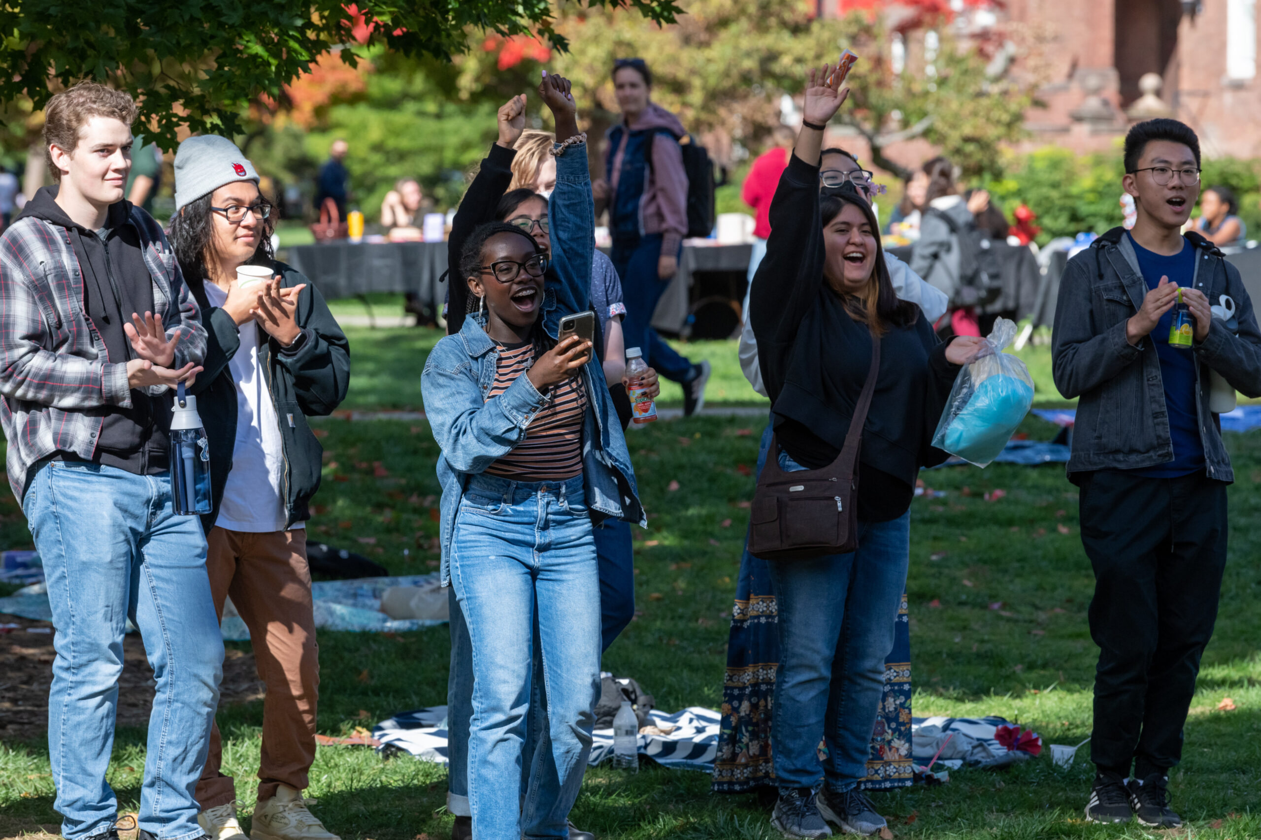 Students standing in the grass clapping and raising their arm in the air as they watch a performance.