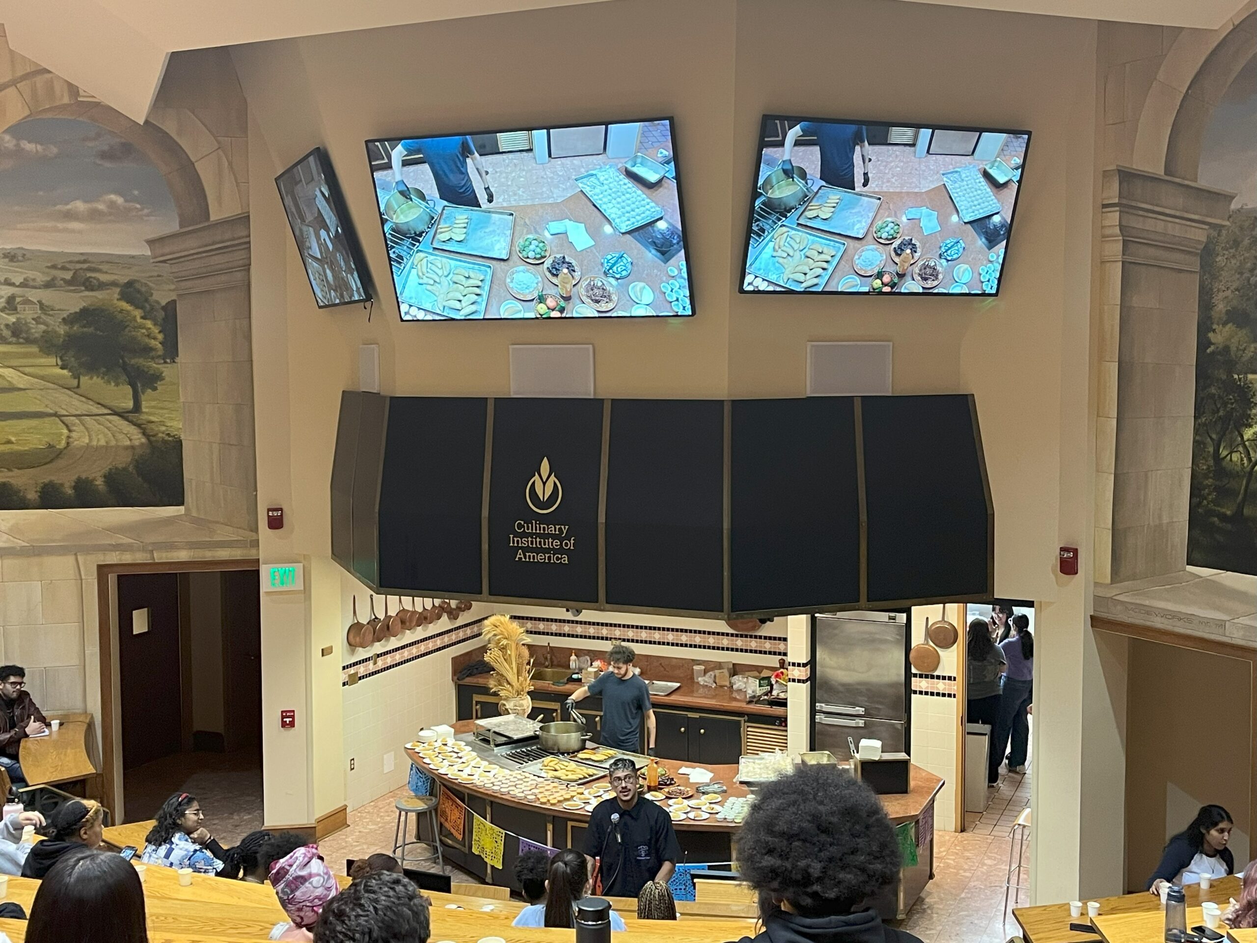 Image of the cooking demo event hosted on CIA Hyde Park's campus. Three screens are pictured with Chef David standing behind the podium speaking into the mic as student are seated lecture styled. 
