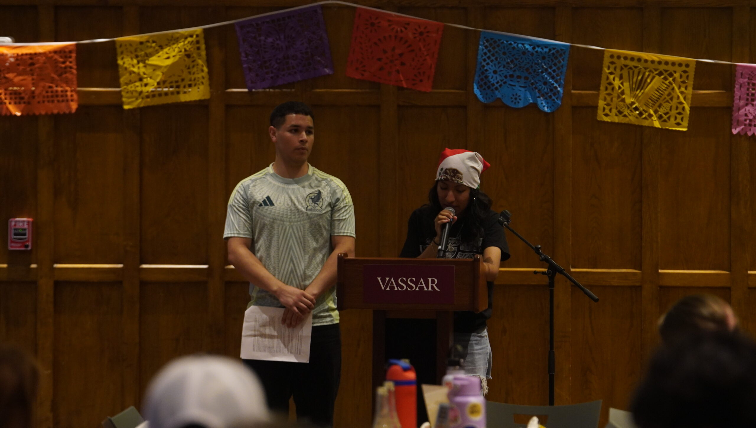 Two student leaders from LSU standing up front behind a podium for the Latine Heritage Month Kickoff in the Villard Room. Orange, Yellow, Red, Blue, Yellow papel picado is hanging on the wooden wall behind them as one student speaks into the mic and the other student standing to the left is crossign their hands in front holding a paper.