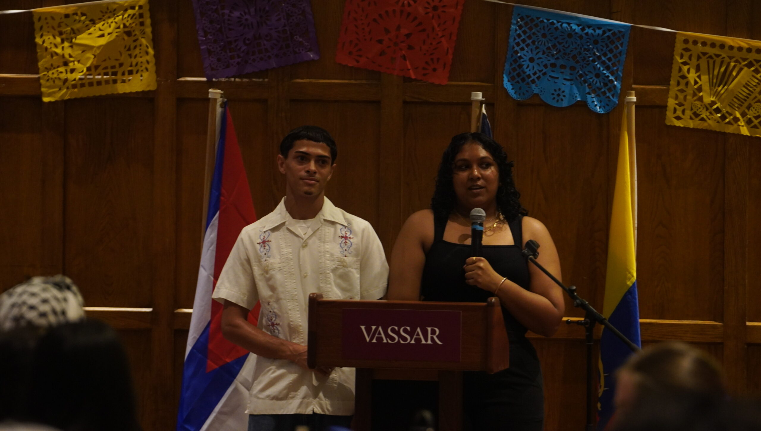 Two student leaders from SOCA standing behind the podium. The student on the left is wearing a white shirt looking out to the audience as the second student is speaking into the mic and wearing a black outfit. 
