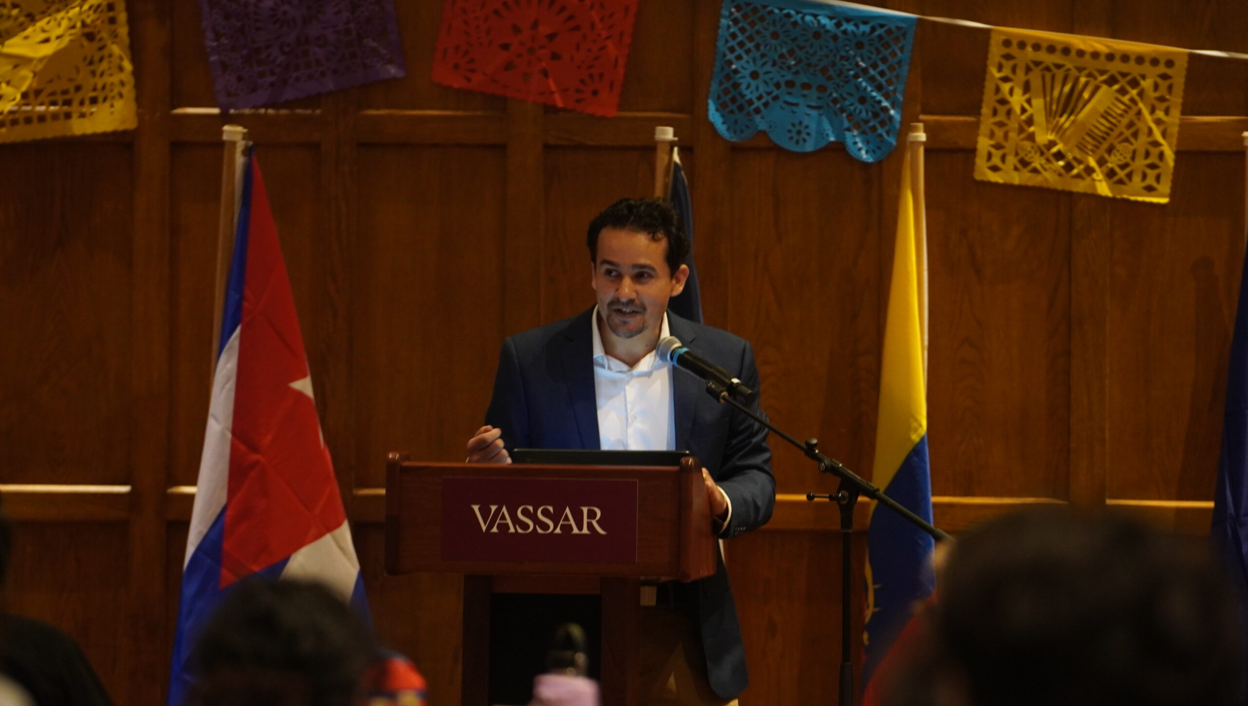 Keynote faculty speaker standing at the podium with the Cuban and Columbian flag to his left and right as he speaks into the mic.
