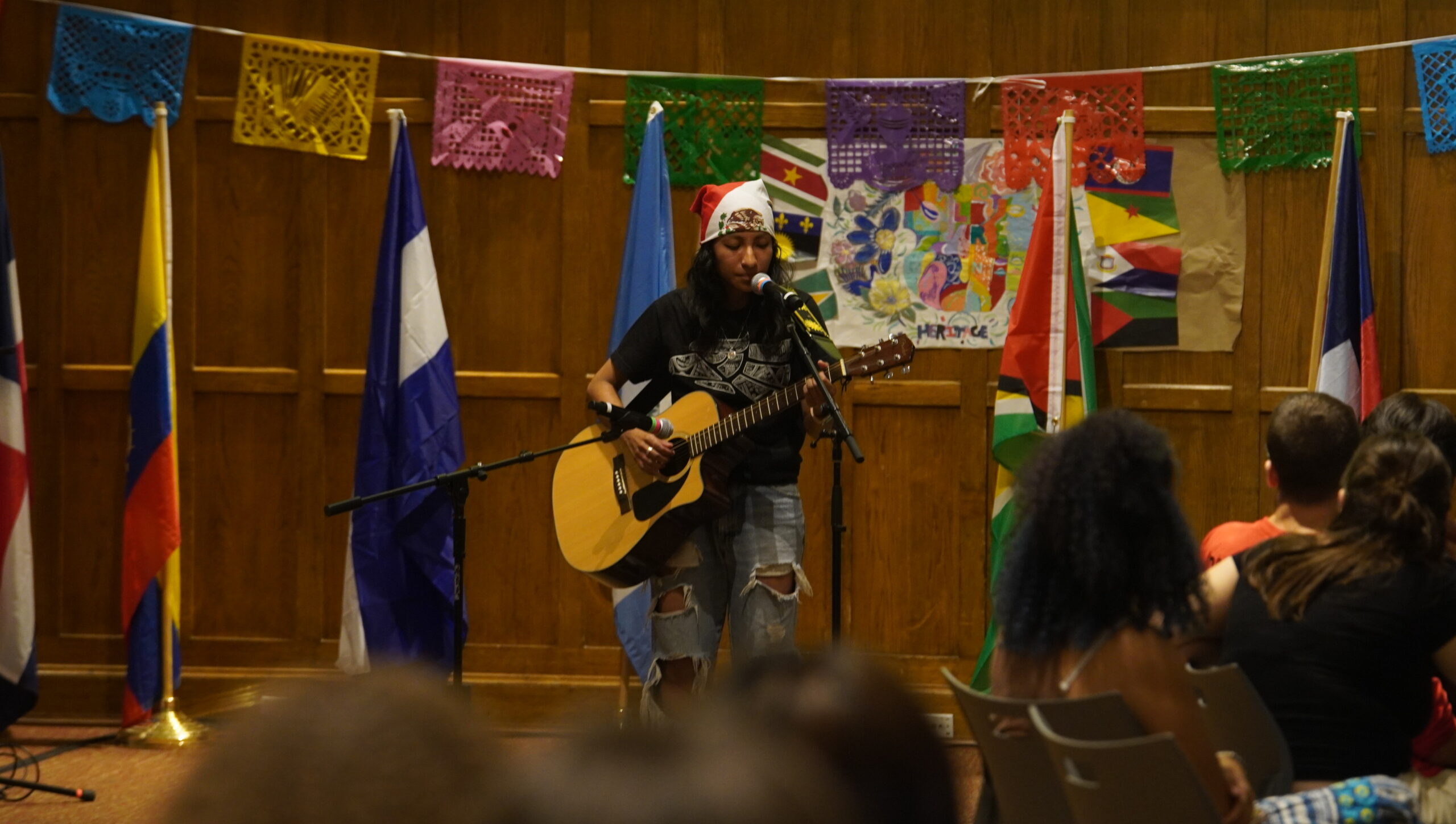 Student leader, standing in front of the audience holding an acoustic guitar and singing into the mic.